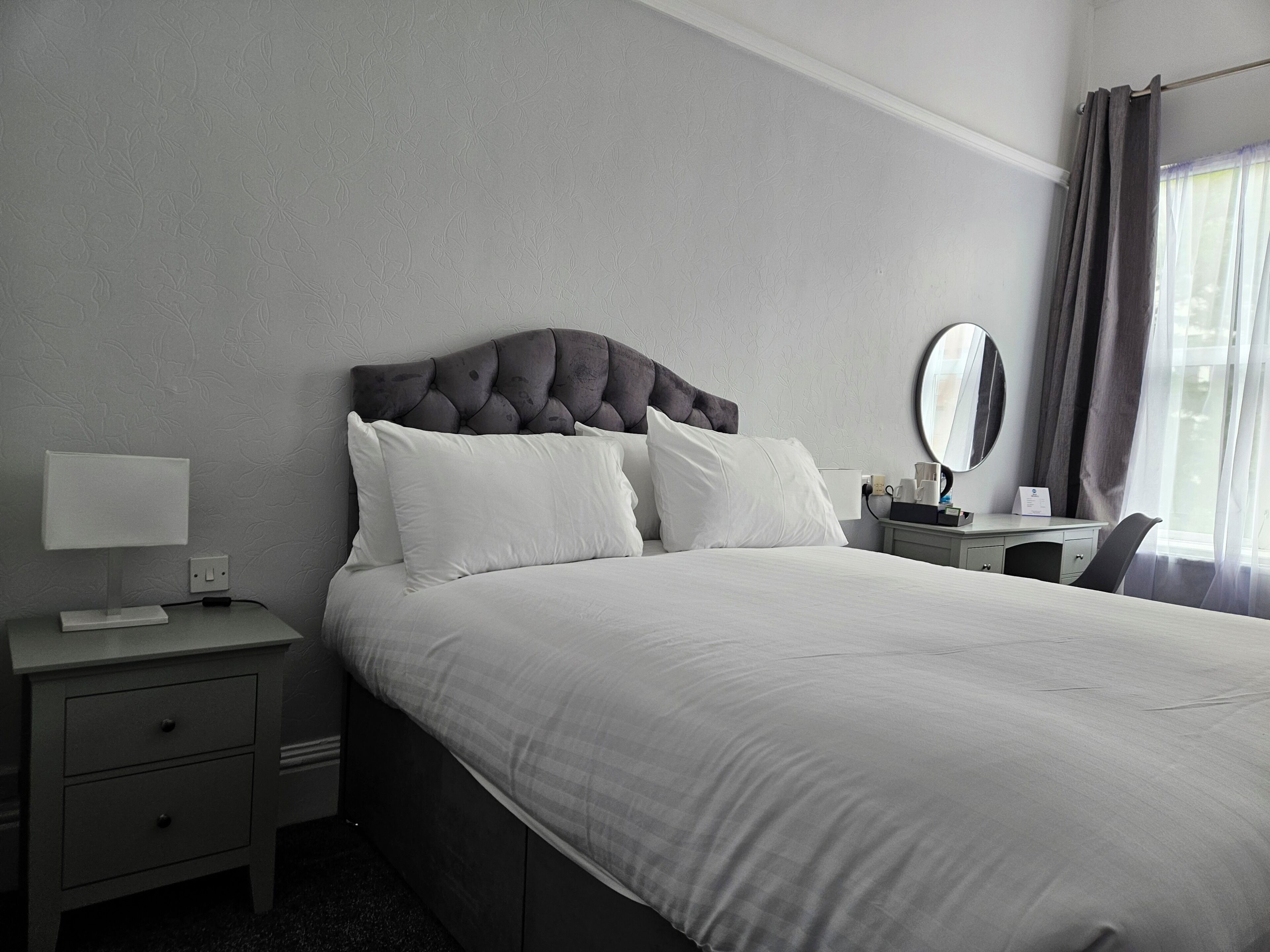 Una habitación de hotel con cama, mesita de noche, lámpara, espejo y escritorio con silla.