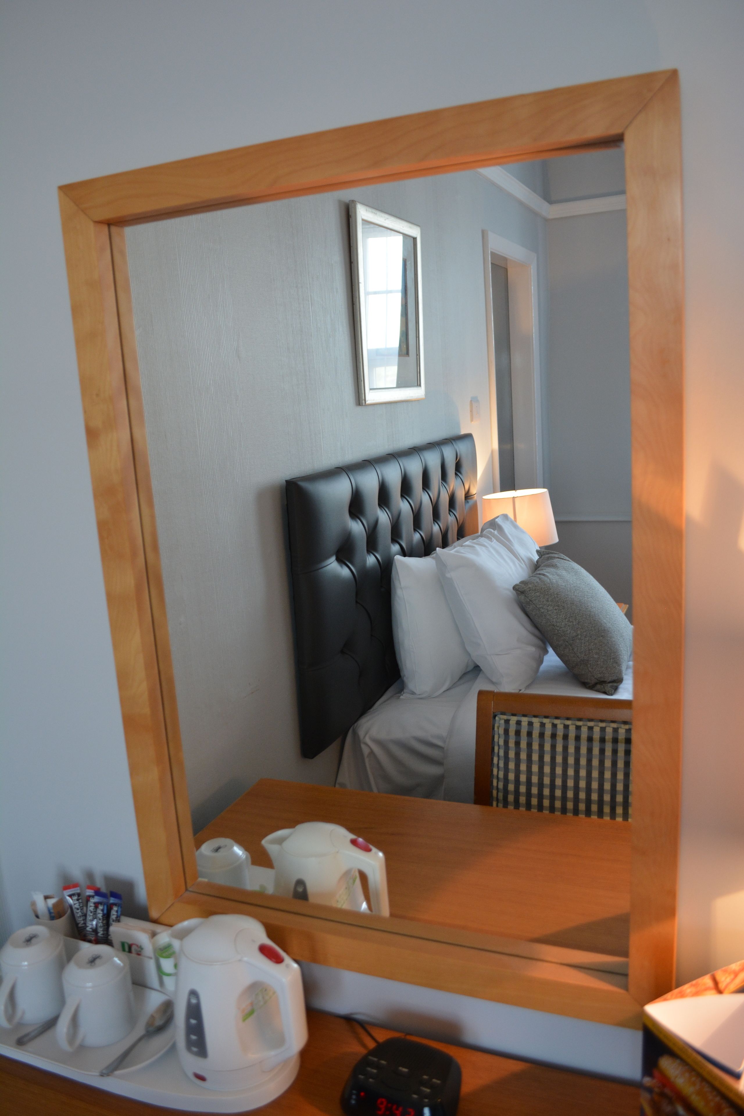 A hotel room with a large mirror reflecting a bed with a black headboard, pillows, and a bedside table with a lamp and a remote control.
