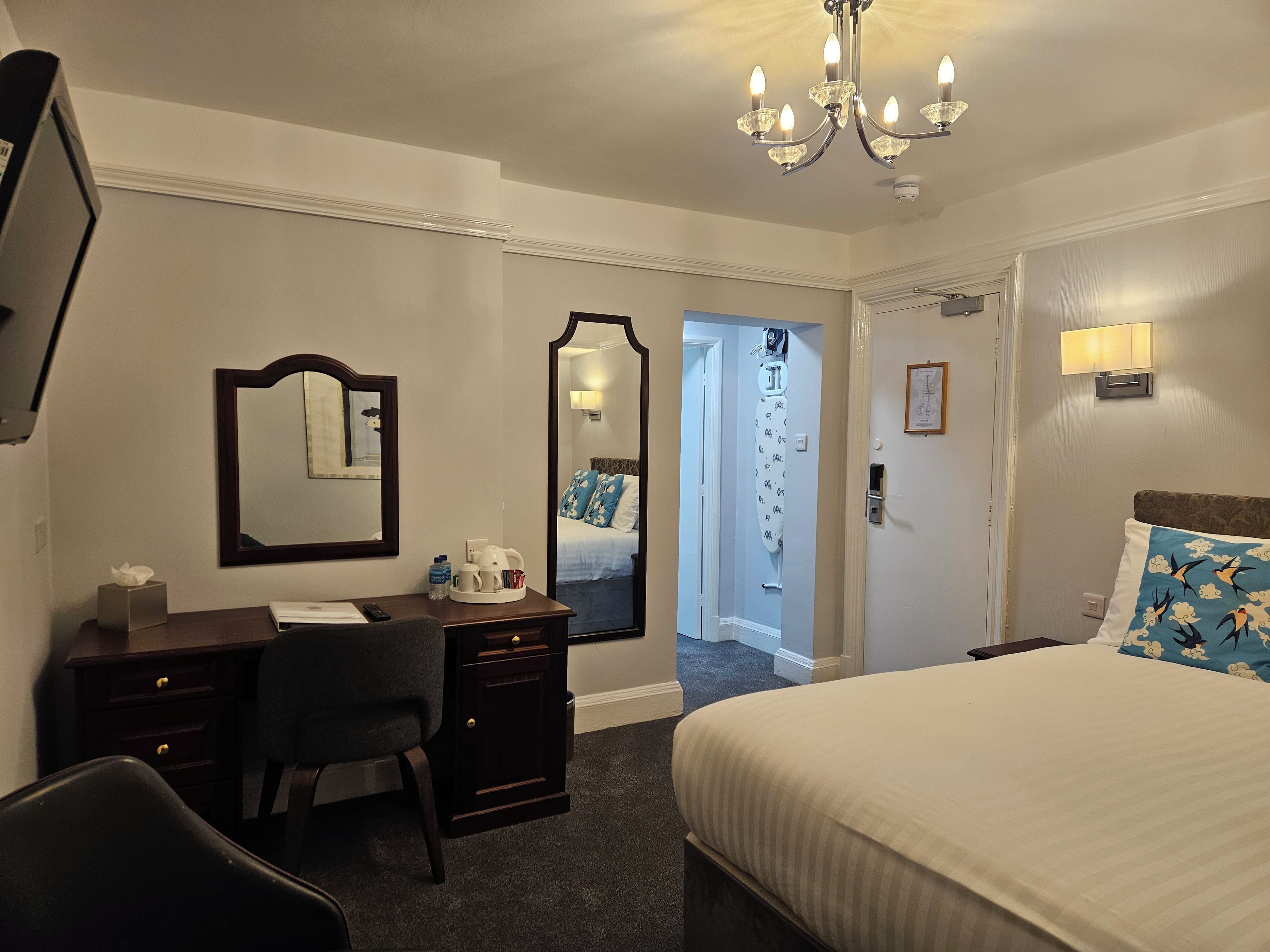 Habitación de hotel con cama, escritorio con silla, espejo, candelabro y puerta que da a otra habitación.