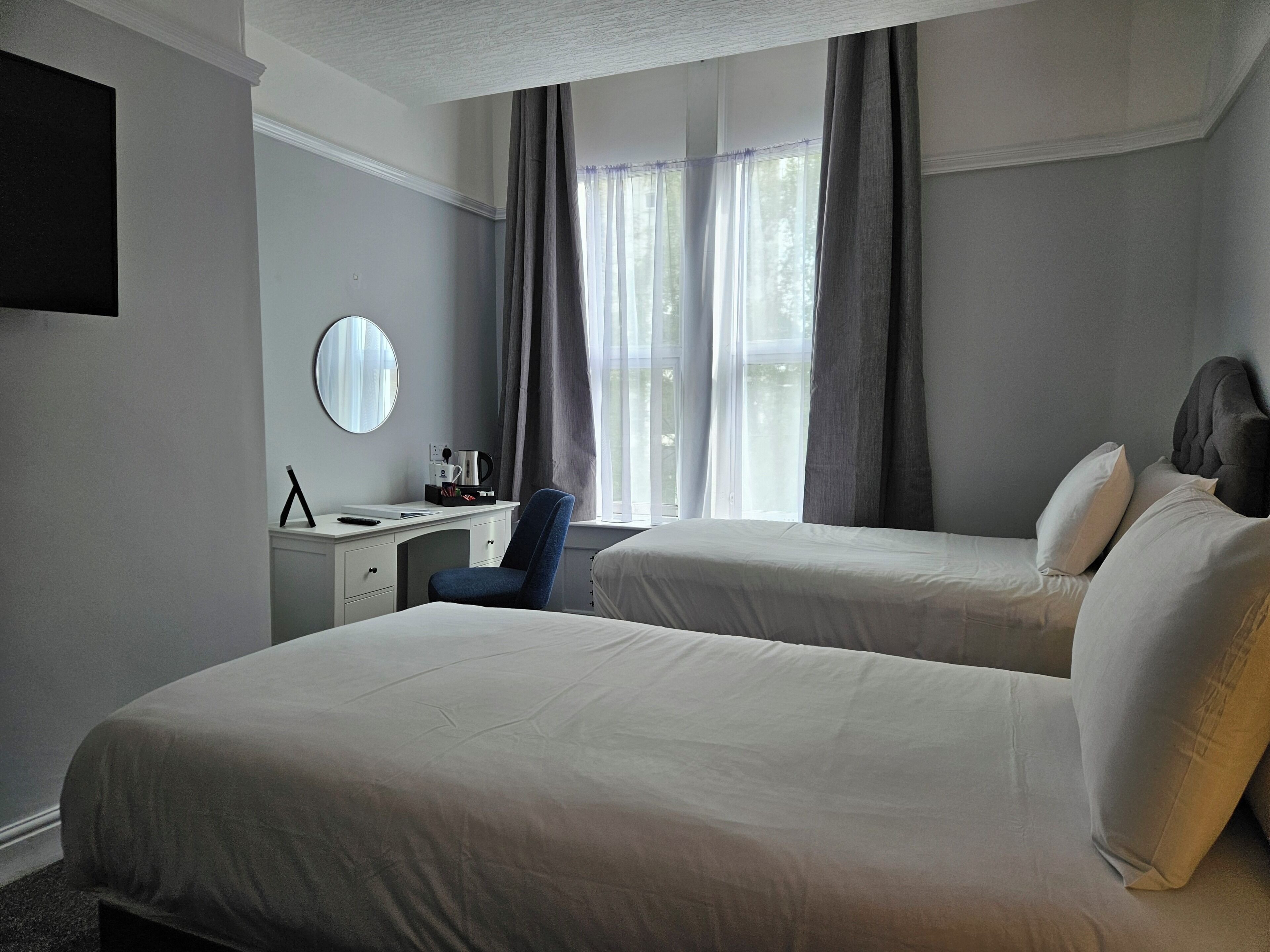 A hotel room with two beds, a desk, a chair, a round mirror, and a window with curtains.