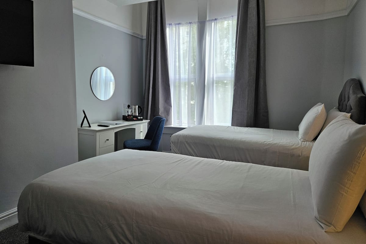 A hotel room with two beds, a desk, a chair, a round mirror, and a window with curtains.