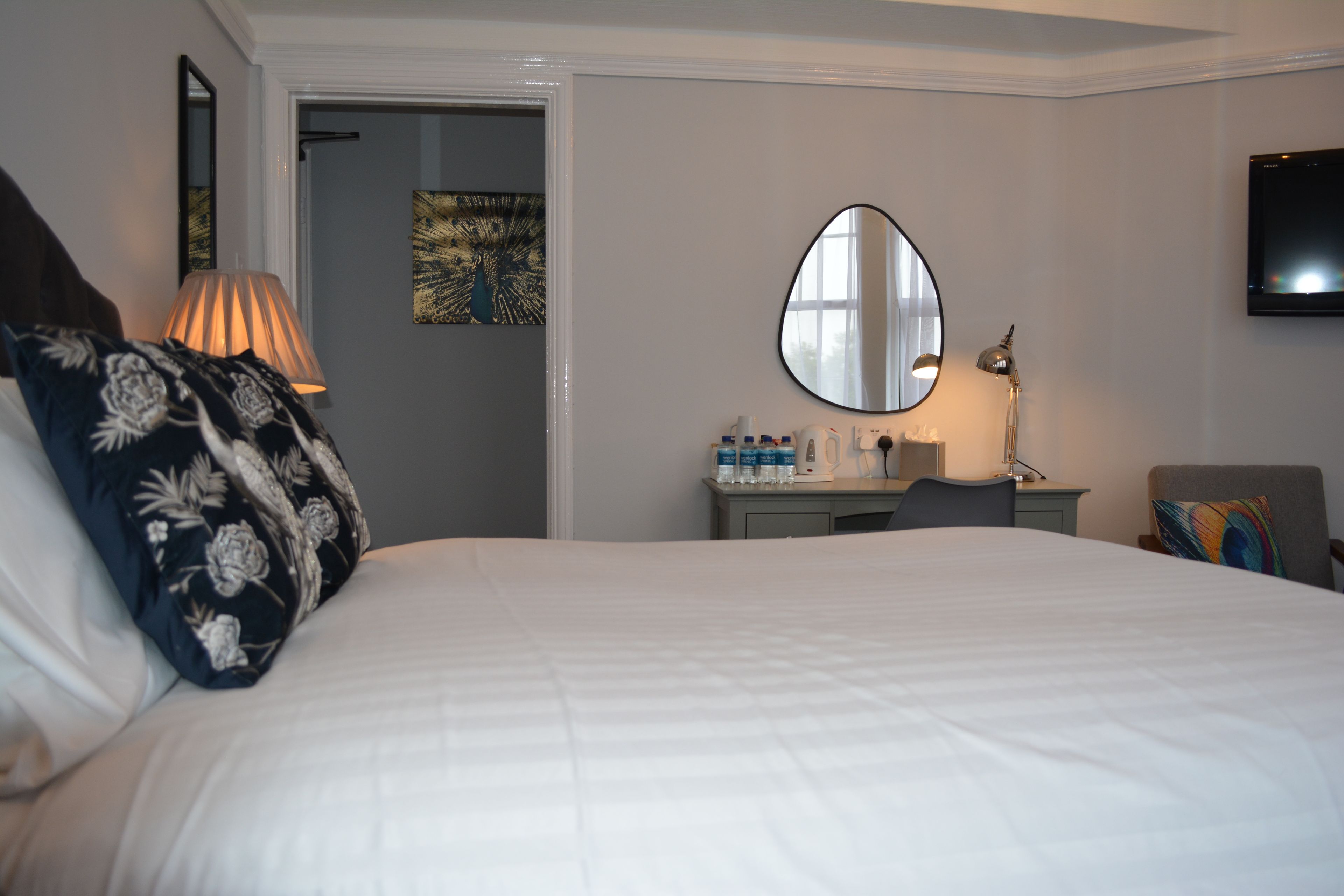 A hotel room with a large bed, a bedside table with a lamp, a mirror, a desk with a chair, and a television on the wall.