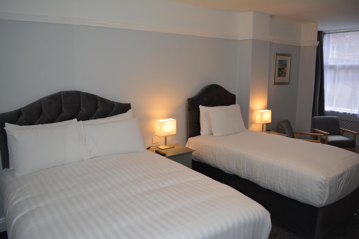 Habitación de hotel con dos camas, un escritorio, una silla y un ventanal con cortinas.