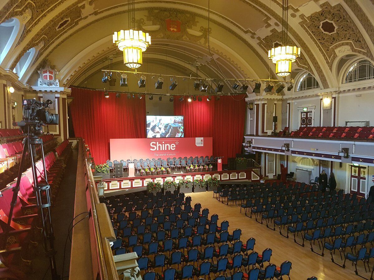 A theater with a red stage, rows of blue chairs, and ornate ceiling details.