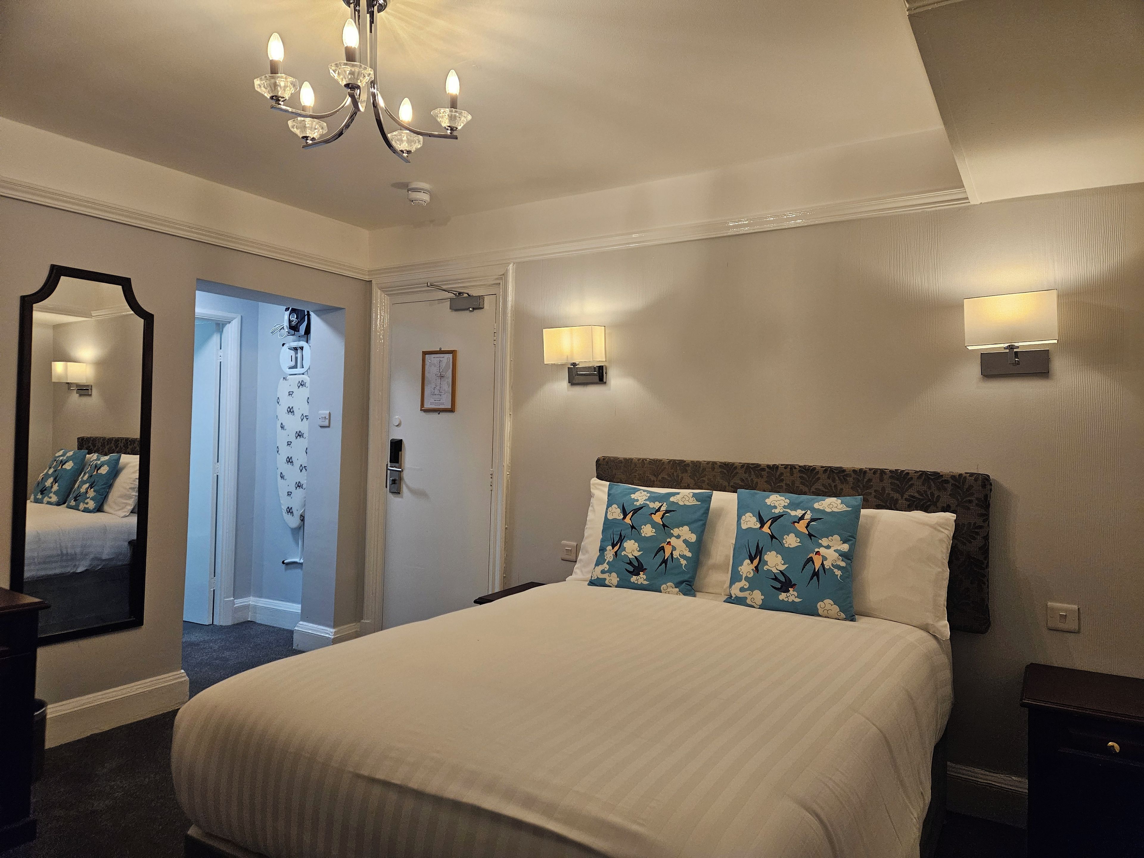 A hotel room with a large bed, a chandelier, a door with a mirror, and wall-mounted lights.