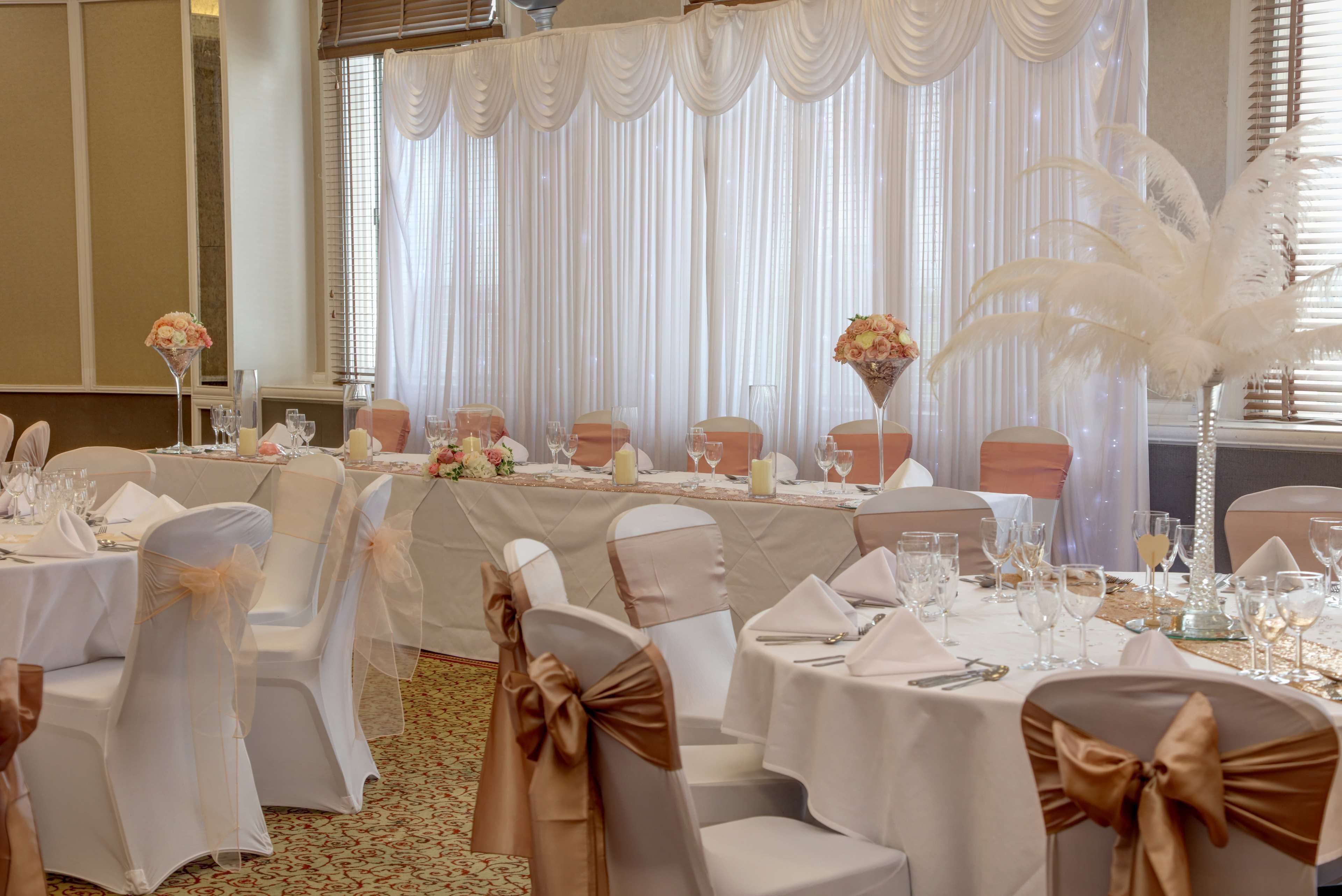 Een balzaal met ronde tafels, witte en gouden stoelen en een witte gordijnenachtergrond.