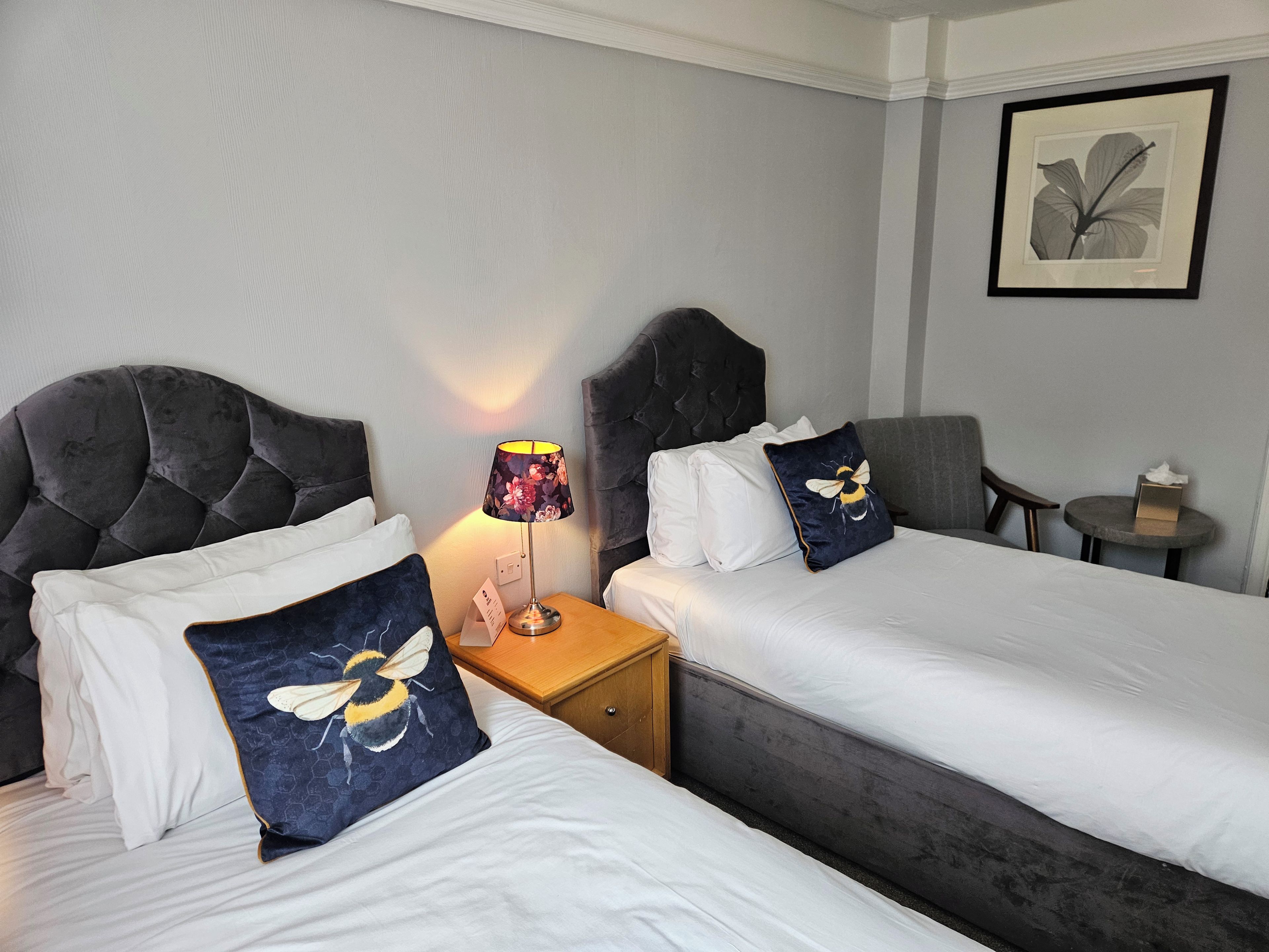 Ein Hotelzimmer mit zwei Betten, jeweils mit dunklem Kopfteil und weißer Bettdecke. Es gibt einen Nachttisch mit einer Lampe und einen kleinen Tisch mit einer Taschentuchbox.