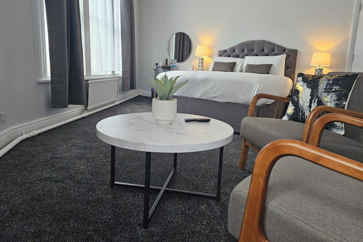 Hotellihuone, jossa on sänky, pyöreä pöytä, jossa on kasvi, kaksi tuolia, lamppu ja ikkuna, jossa on verhot.