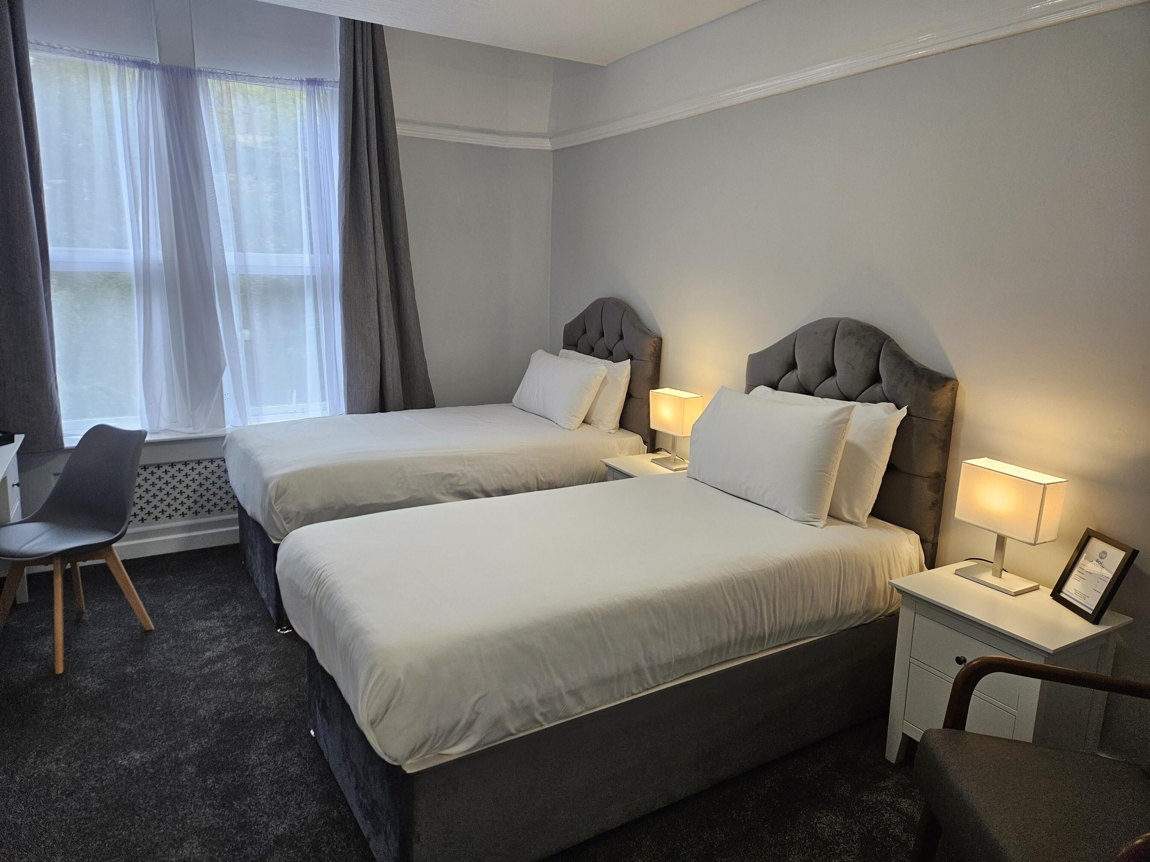 A hotel room with two beds, a desk, a chair, a window with curtains, and a bedside table with a lamp and a framed certificate.
