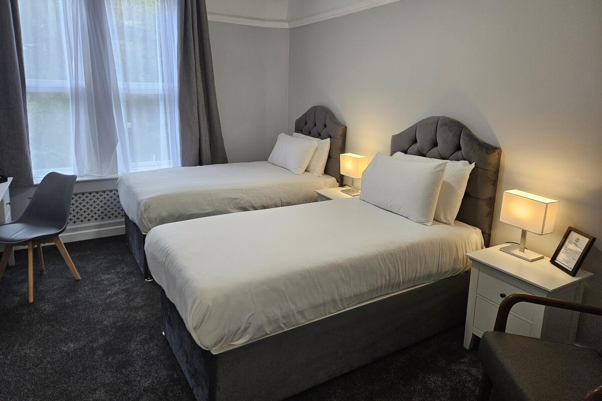 Habitación de hotel con dos camas, un escritorio, una silla, un ventanal con cortinas y una mesita de noche con una lámpara y un diploma enmarcado.