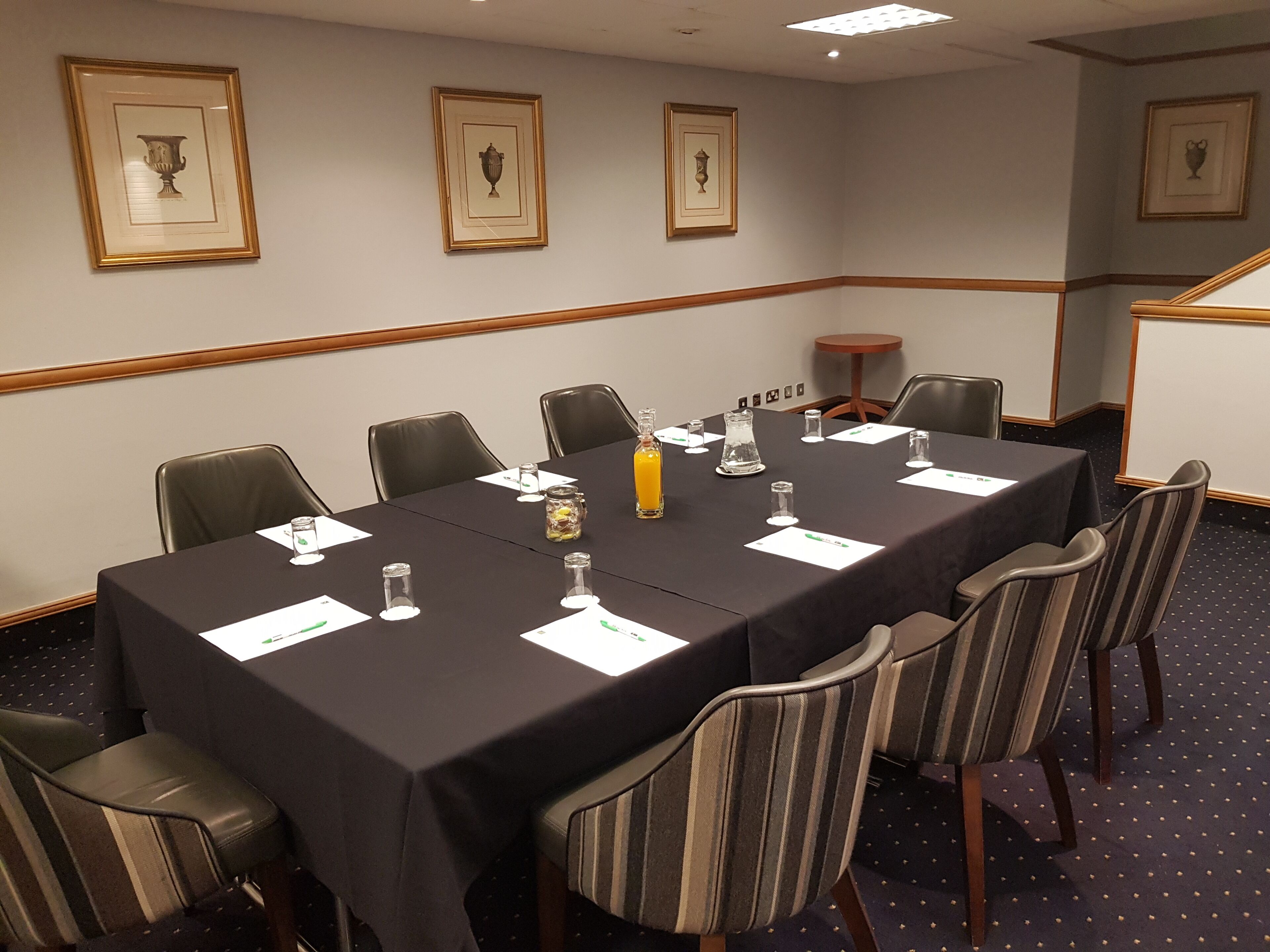 Un salón de conferencias con una larga mesa, sillas, cuadros en la pared y una pequeña mesa con una botella y vasos.