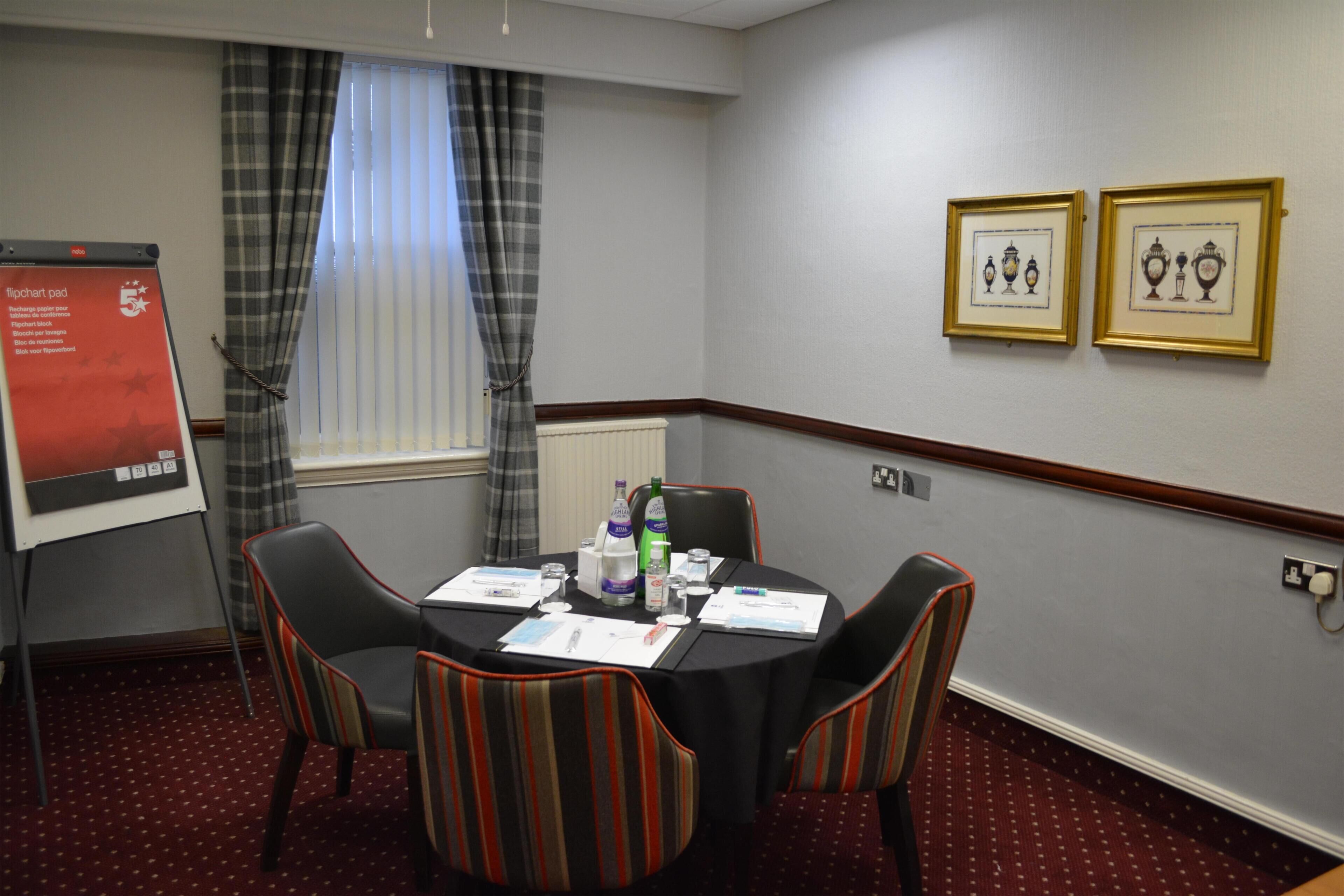 A conference room with a round table, four striped chairs, a flipchart pad, and two framed pictures on the wall.