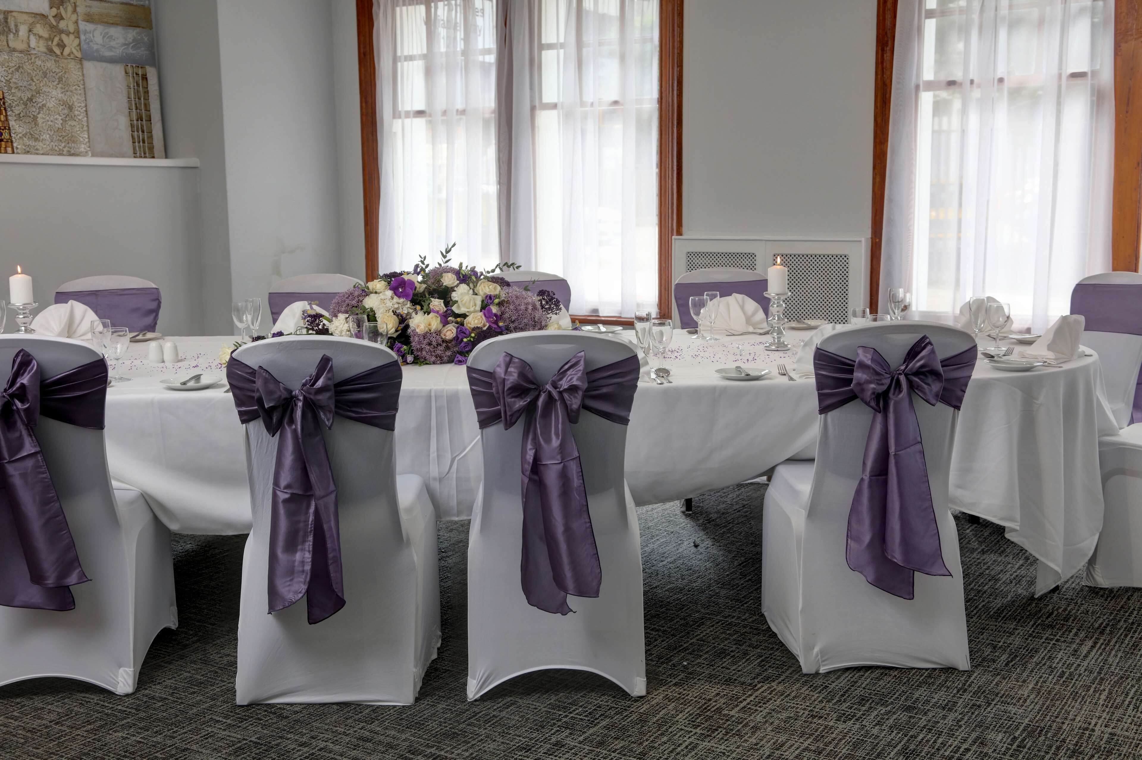 Un salón de banquetes con mesas redondas, manteles blancos y cintas púrpuras en las sillas.