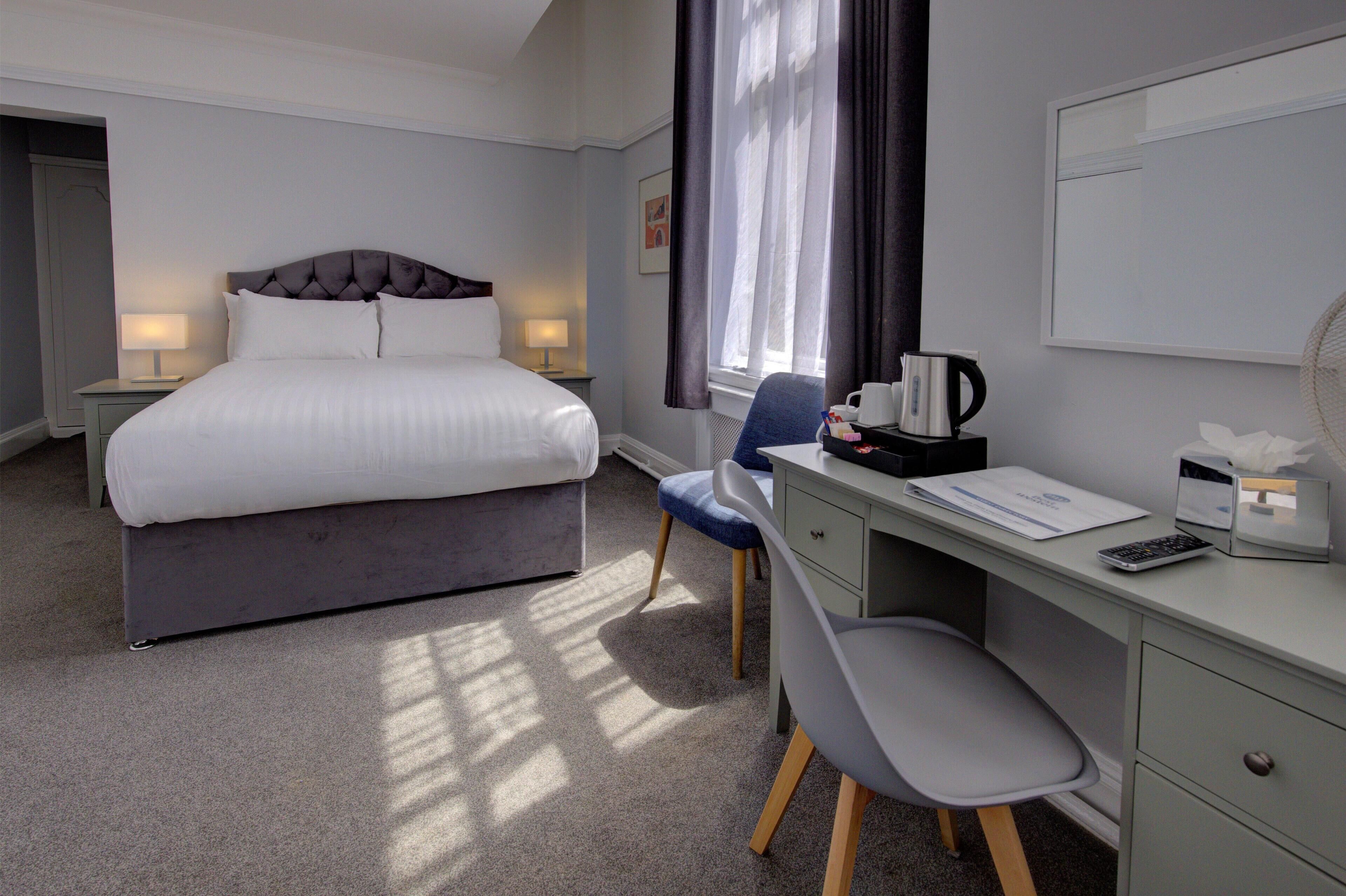 A hotel room with a bed, bedside lamps, a desk with a chair, a coffee maker, and a window with curtains.