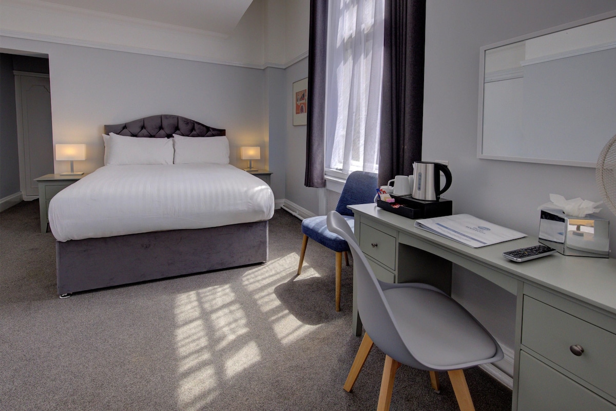 Una habitación de hotel con cama, lámparas de noche, un escritorio con silla, cafetera y ventana con cortinas.