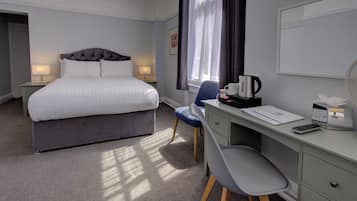 A hotel room with a bed, bedside lamps, a desk with a chair, a coffee maker, and a window with curtains.