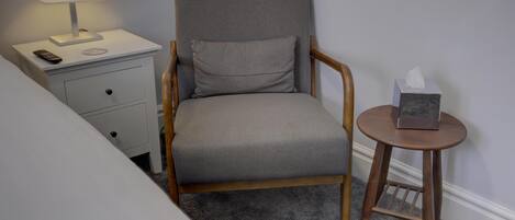 Una butaca tapizada en gris con estructura de madera, una mesita de noche con una lámpara y una pequeña mesa redonda con una caja de pañuelos.