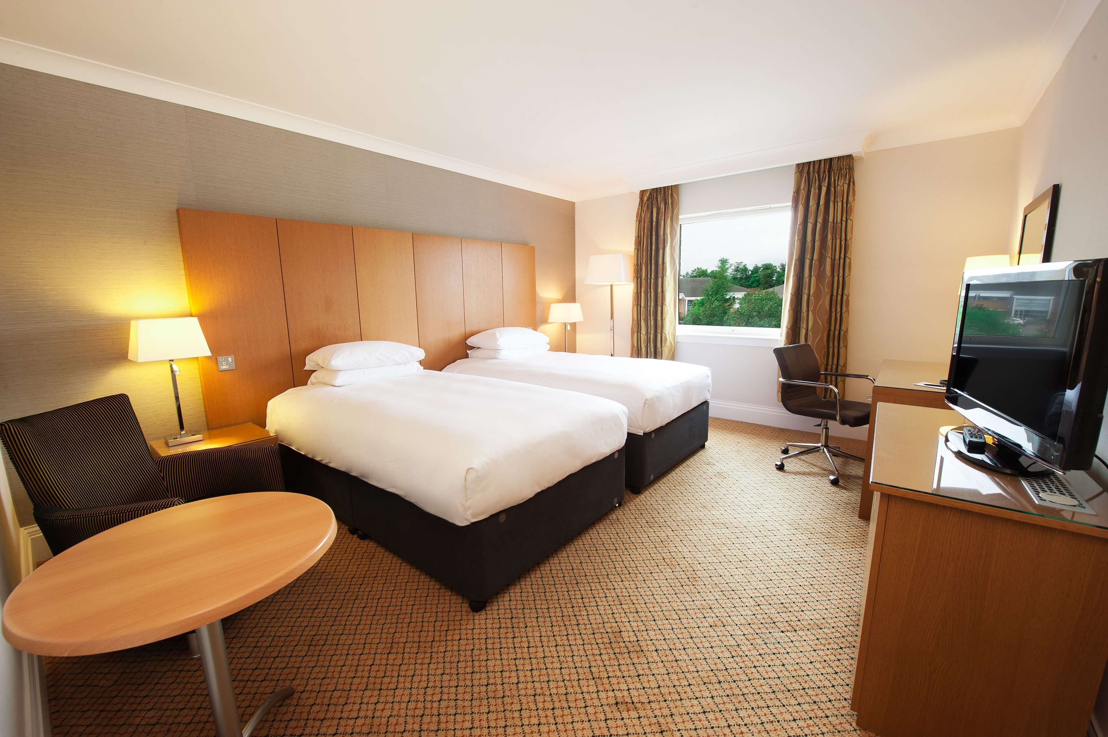 Quarto de hotel com duas camas, uma escrivaninha com computador, uma cadeira, um abajur e uma televisão.