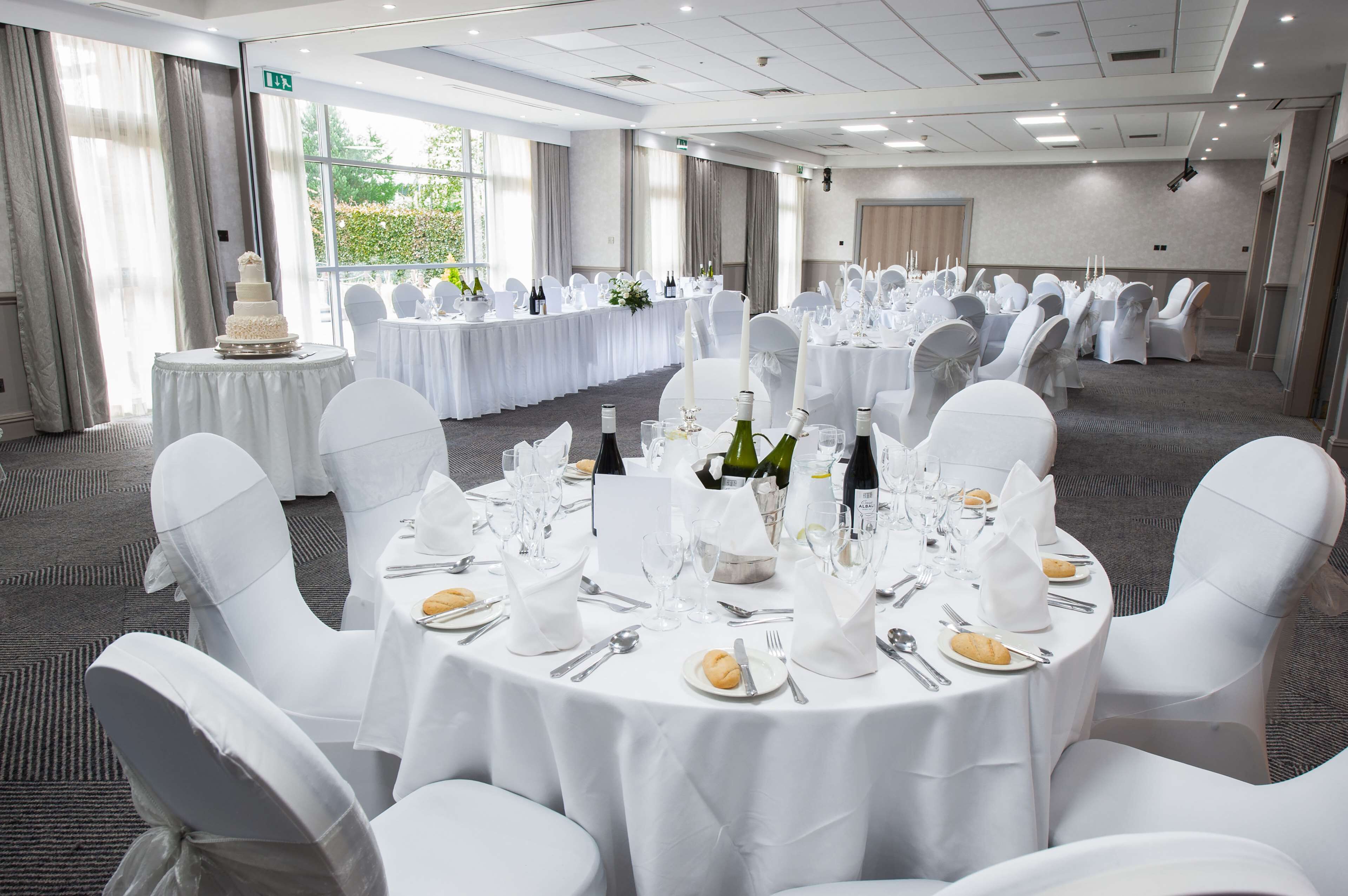 A banquet hall with round tables set for a formal event, featuring white tablecloths, chairs with white covers, and a cake on a pedestal.