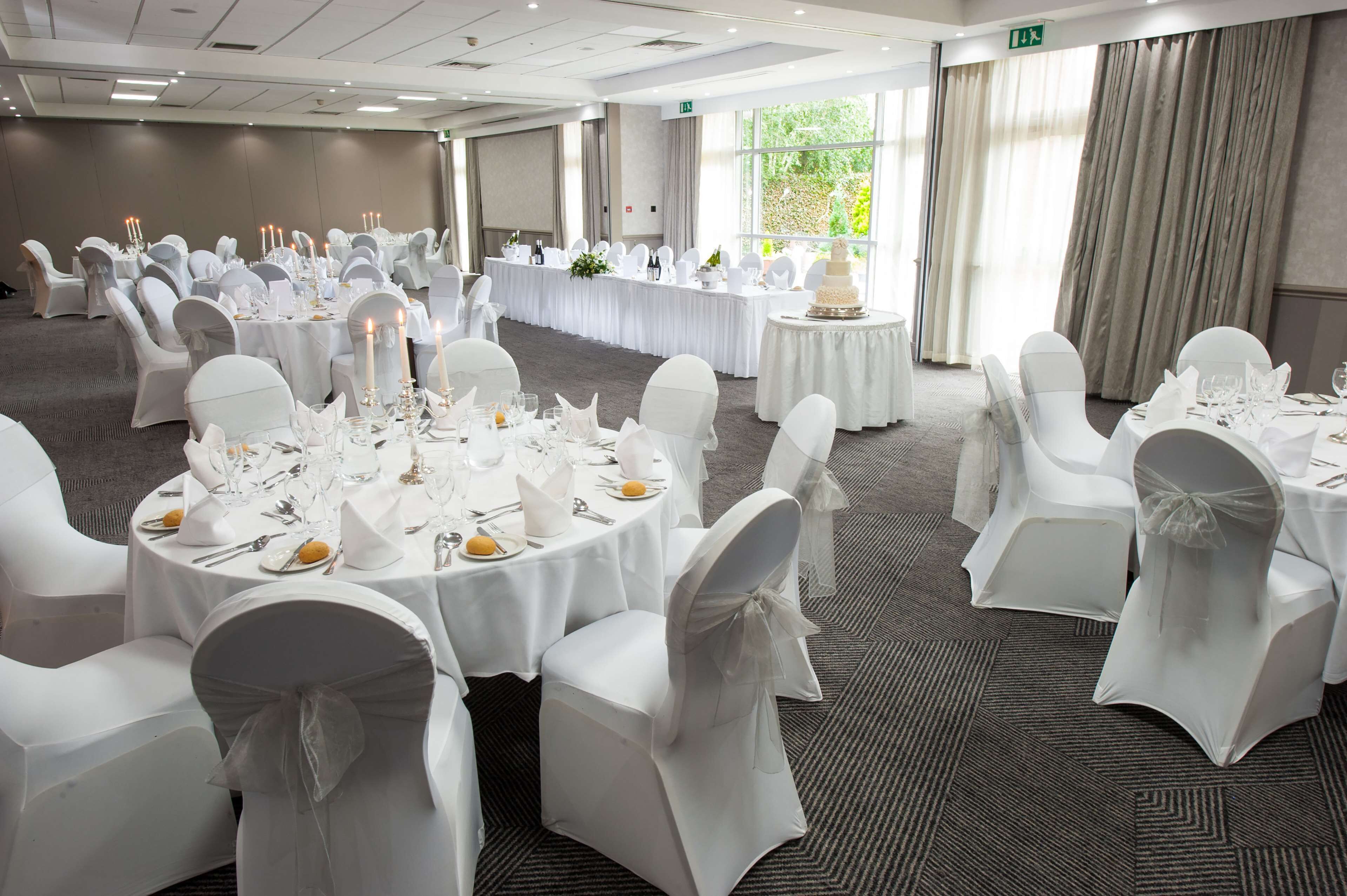 Un salón de banquetes con mesas redondas dispuestas para un evento formal, con manteles blancos, sillas cubiertas de blanco y un gran pastel como pieza central.