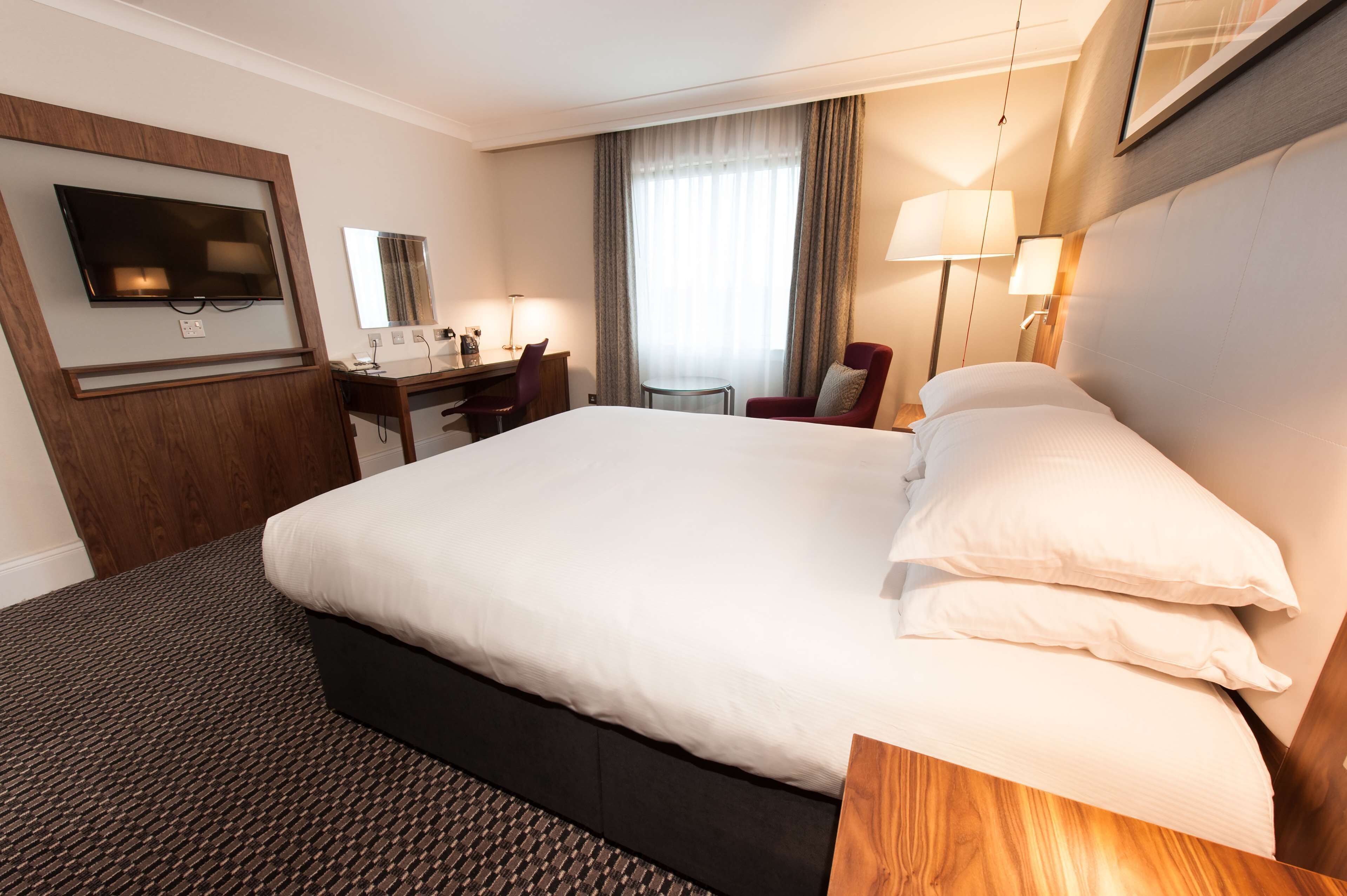 Ein Hotelzimmer mit einem großen Bett, einem Schreibtisch, einem Sessel, einem Fernseher und einem Fenster mit Vorhängen.