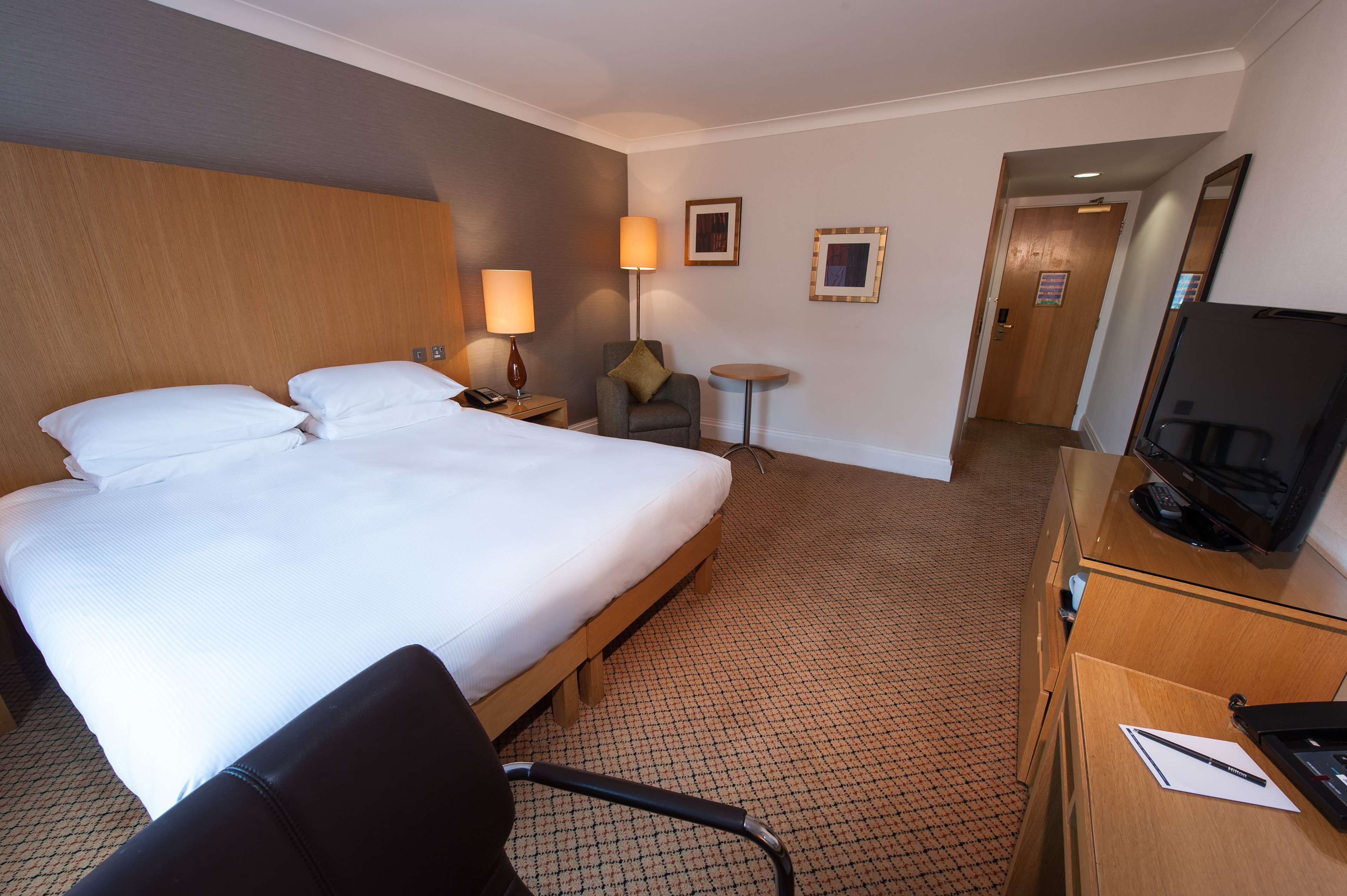 A hotel room with a large bed, a desk with a television, a chair, a small table, and a lamp.