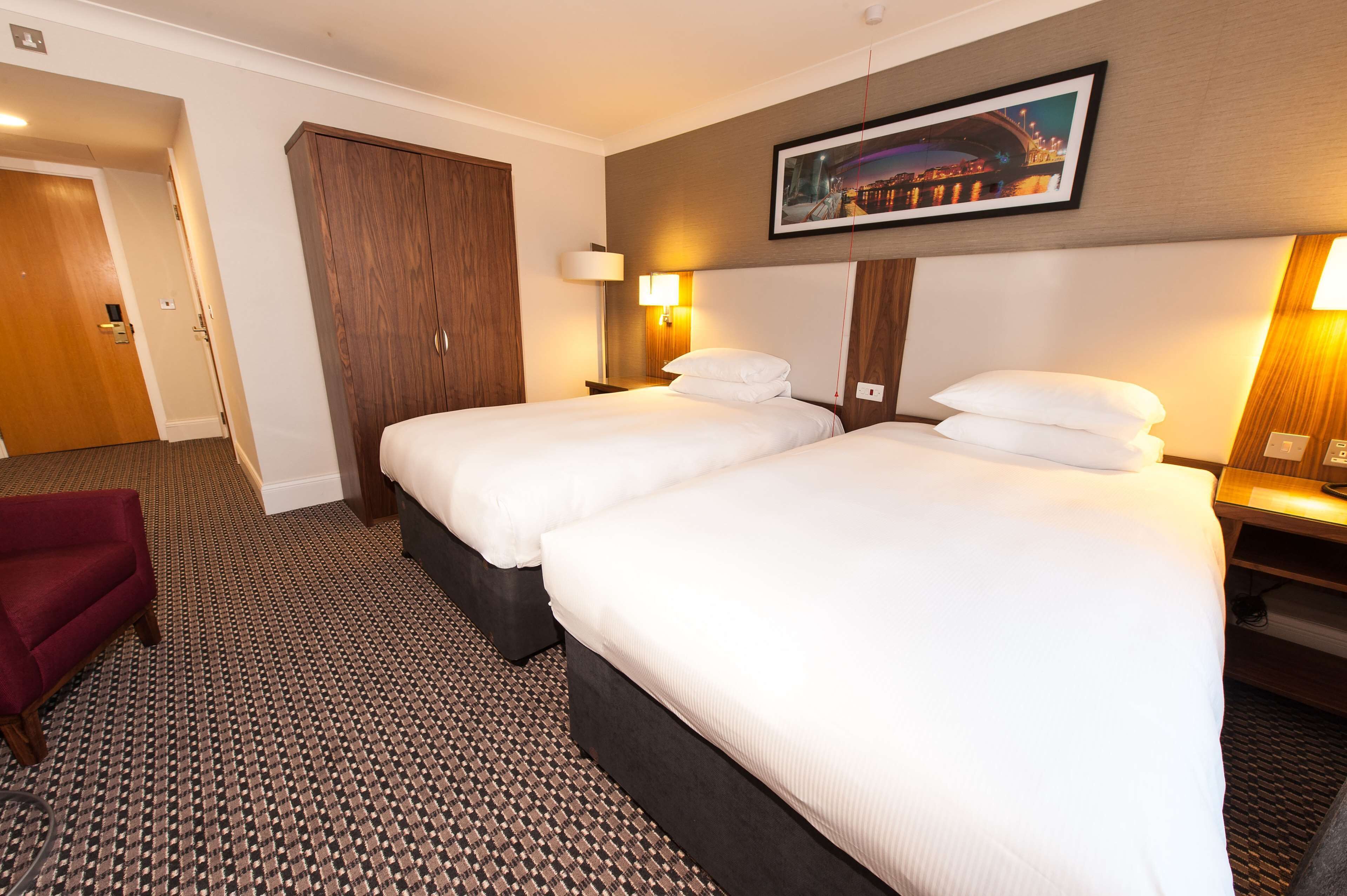 A hotel room with two beds, a wardrobe, a red chair, and a framed picture on the wall.