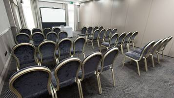 Un salón de conferencias con filas de sillas, una pantalla de proyector y una mesa con un ordenador portátil.
