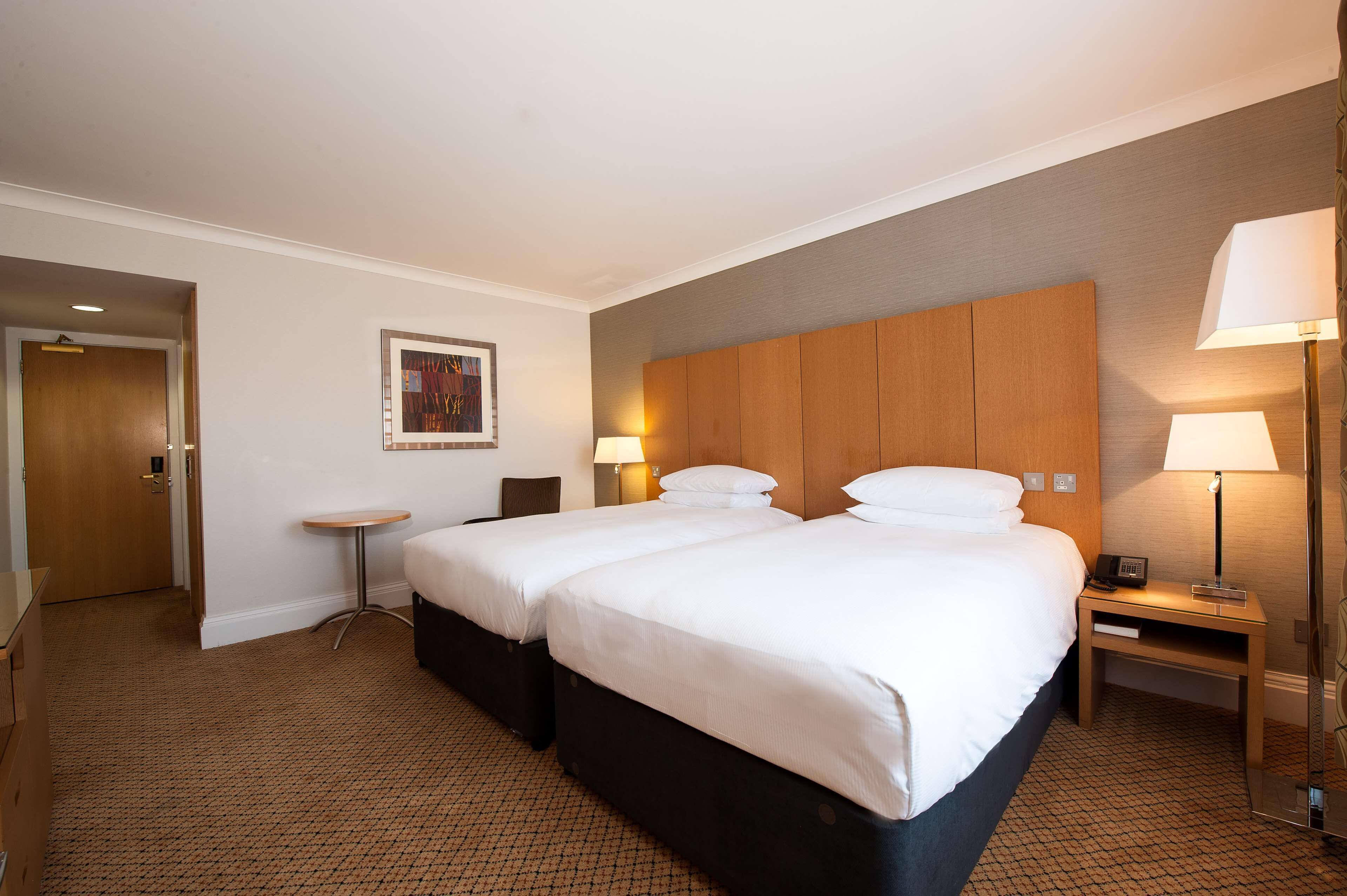 Une chambre d’hôtel avec deux lits, une petite table, une table de chevet avec une lampe et une porte donnant sur une autre pièce.