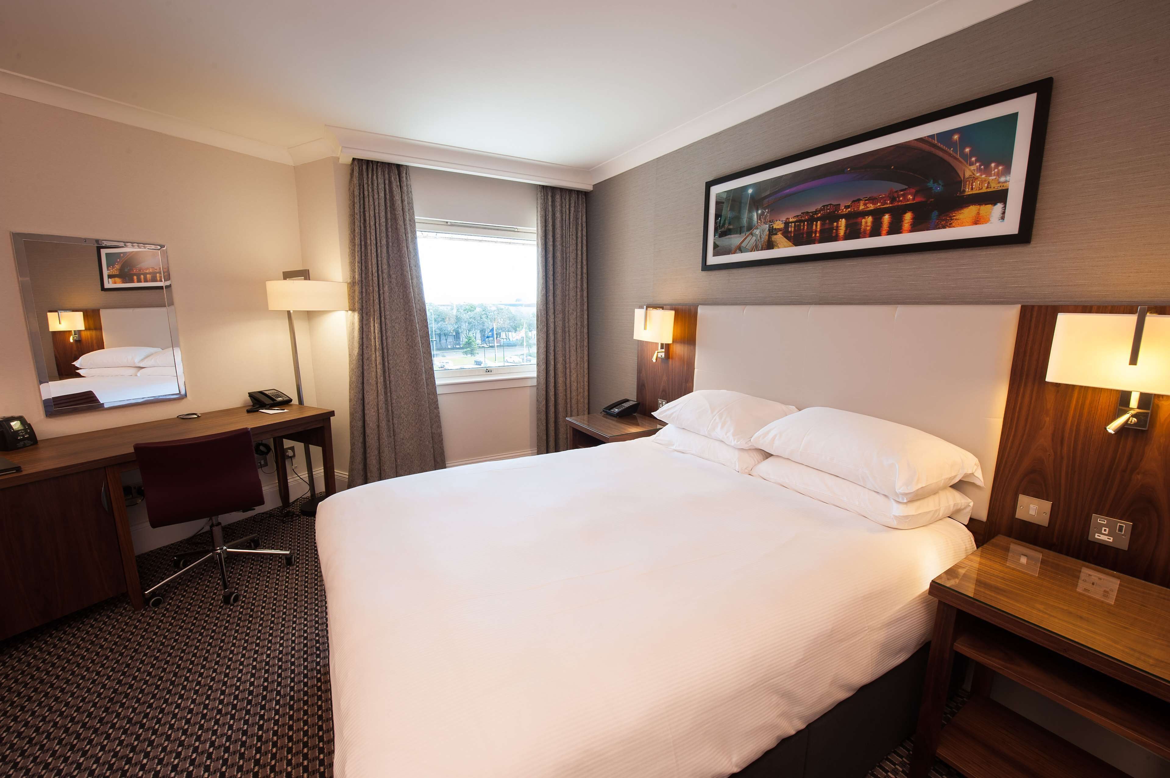 A hotel room with a large bed, a desk with a chair, a desk lamp, a bedside table with a lamp, a window with curtains, and a framed picture on the wall.