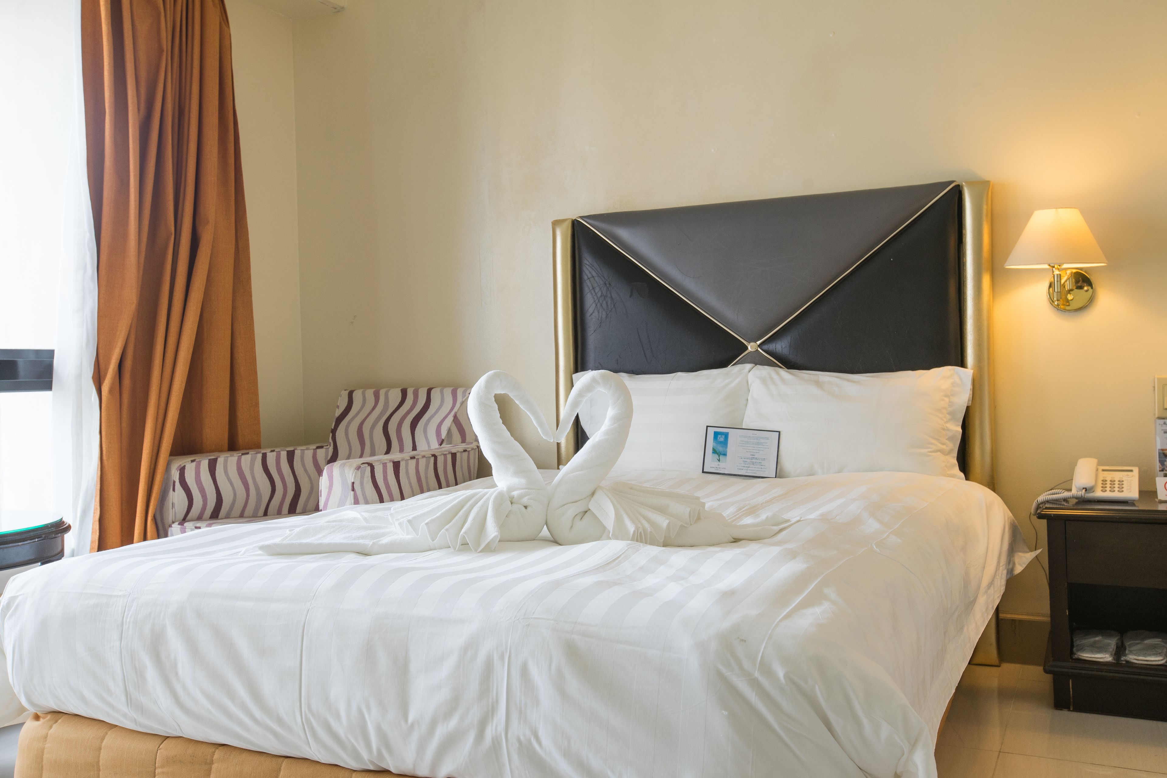 A hotel room with a neatly made bed, a bedside table with a lamp, and a view of the outside through a window with curtains.