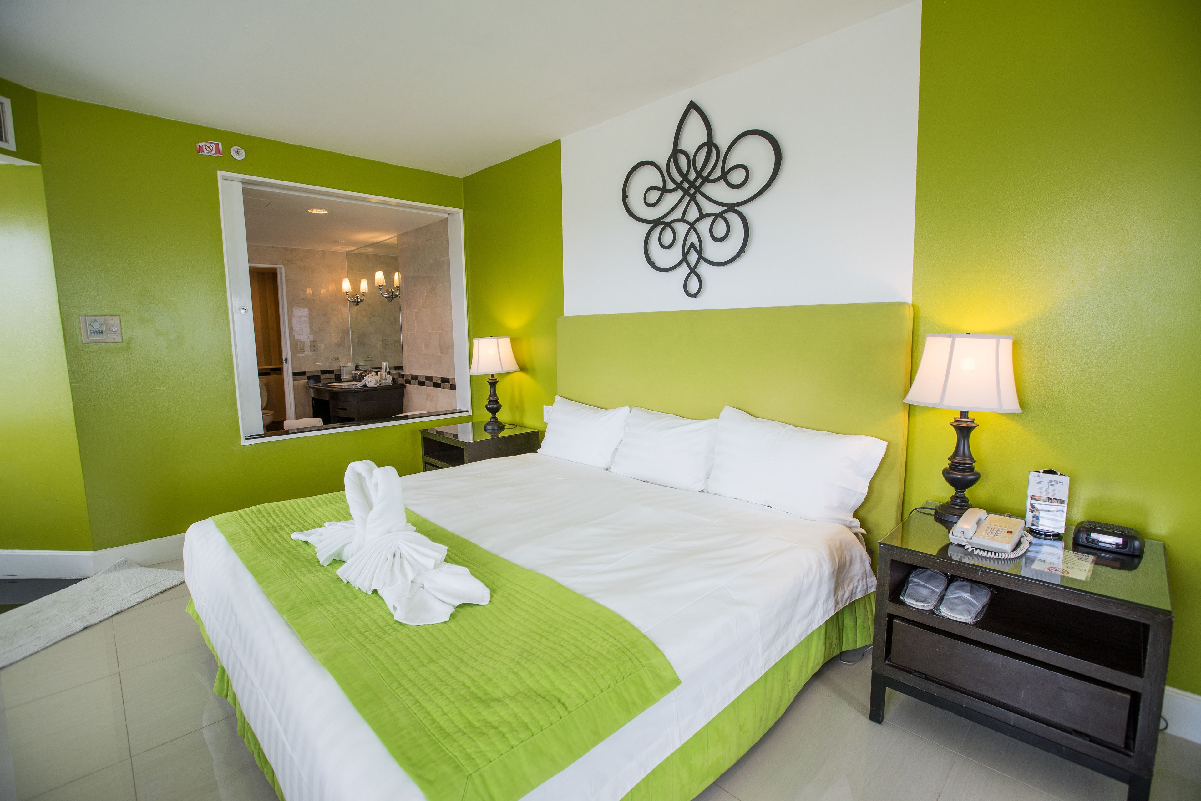 A hotel room with a large bed, green walls, a mirror, two bedside lamps, and a nightstand with a phone and a small table.