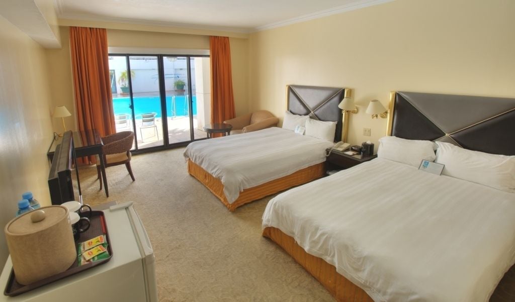 Ein Hotelzimmer mit zwei Betten, einem Schreibtisch mit einem Laptop, einem Stuhl und Blick auf Pool und Strand durch große Fenster.