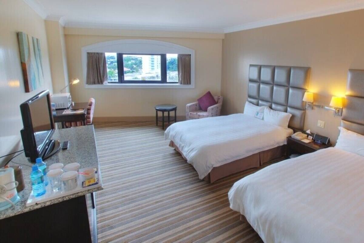 A hotel room with two beds, a desk with a computer, a chair, a small table, and a window with curtains.