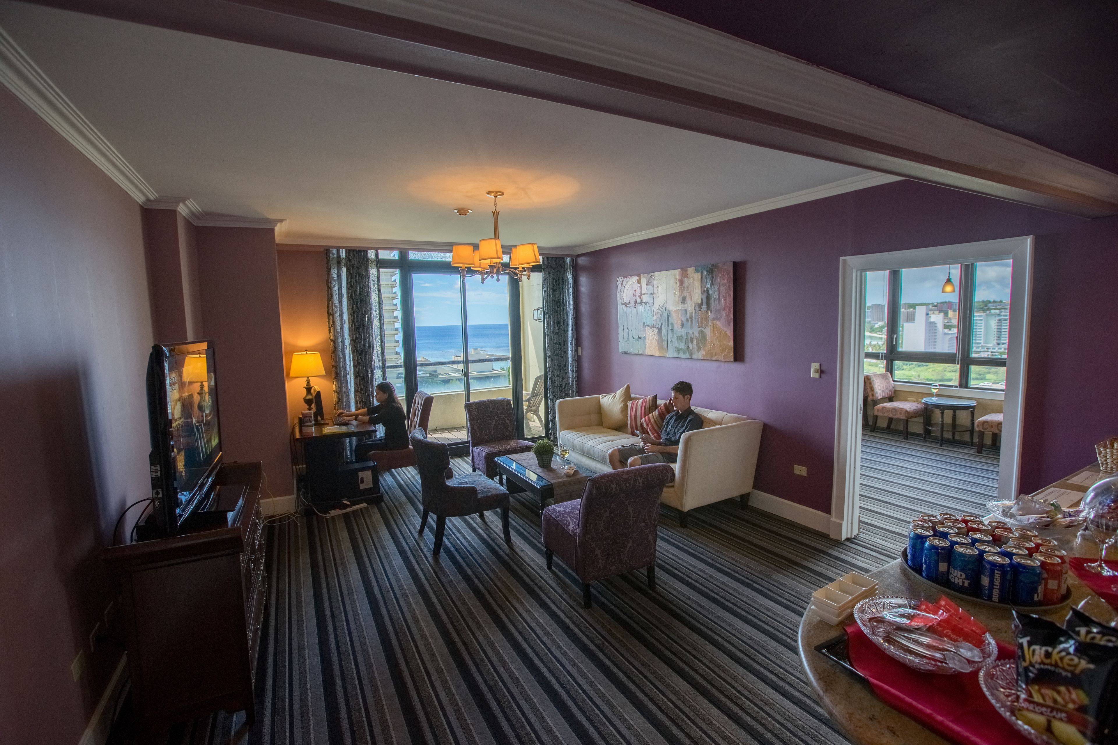 A hotel room with a view of the ocean, a dining area with snacks, and a seating area with a chandelier.