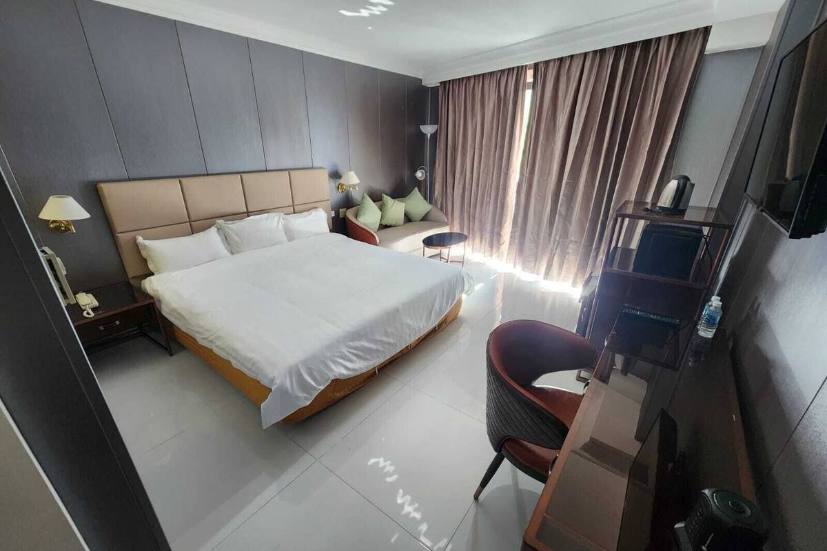 A hotel room with a large bed, a desk with a chair, a TV, and a bedside table with a lamp.