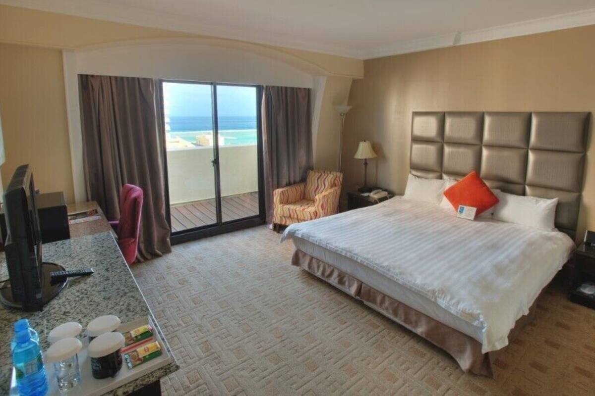 A hotel room with a large bed, a desk with a computer, a chair, and a view of the beach through a sliding door.