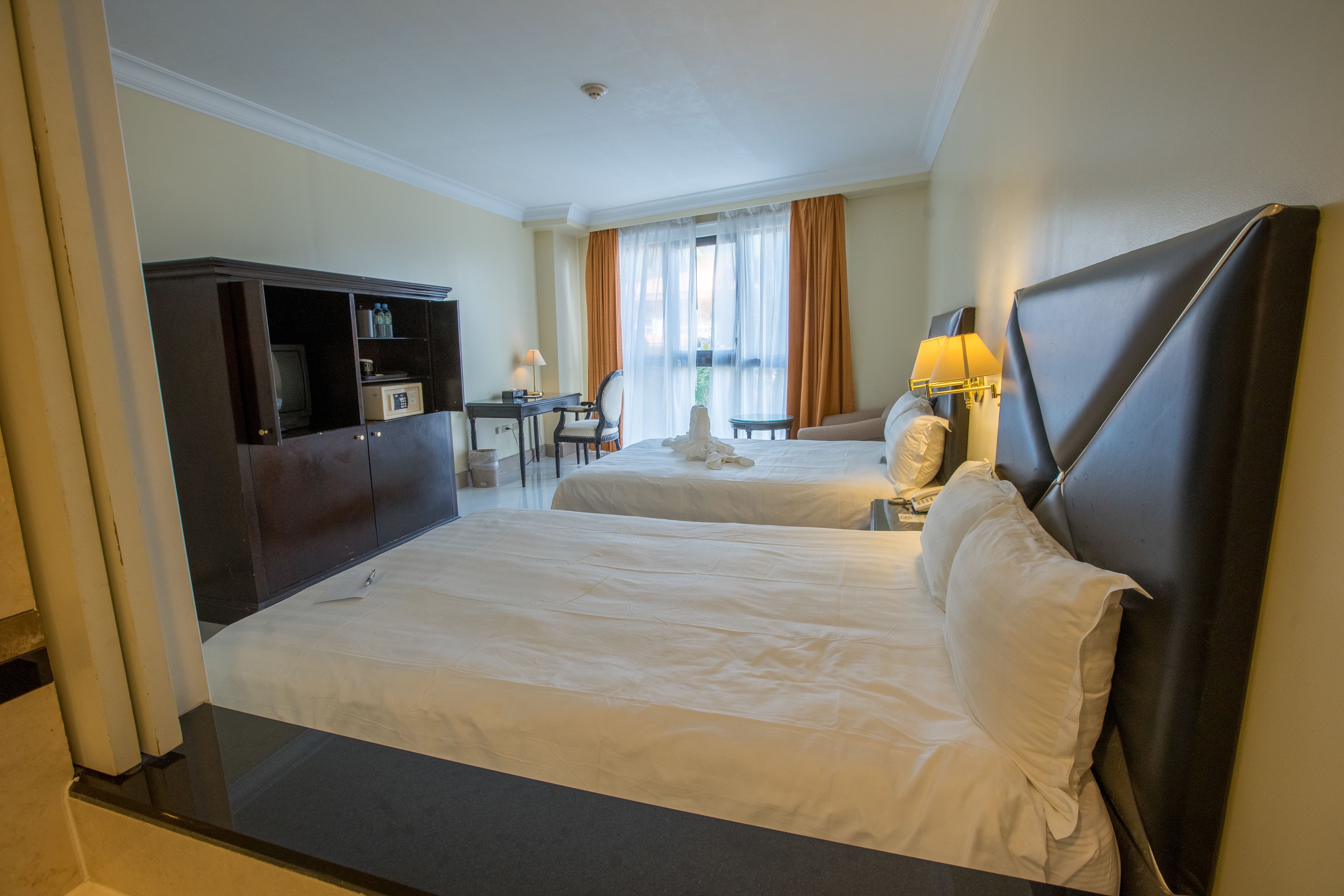 Ett hotellrum med två sängar, ett skrivbord, en stol, en tv och ett fönster med gardiner.