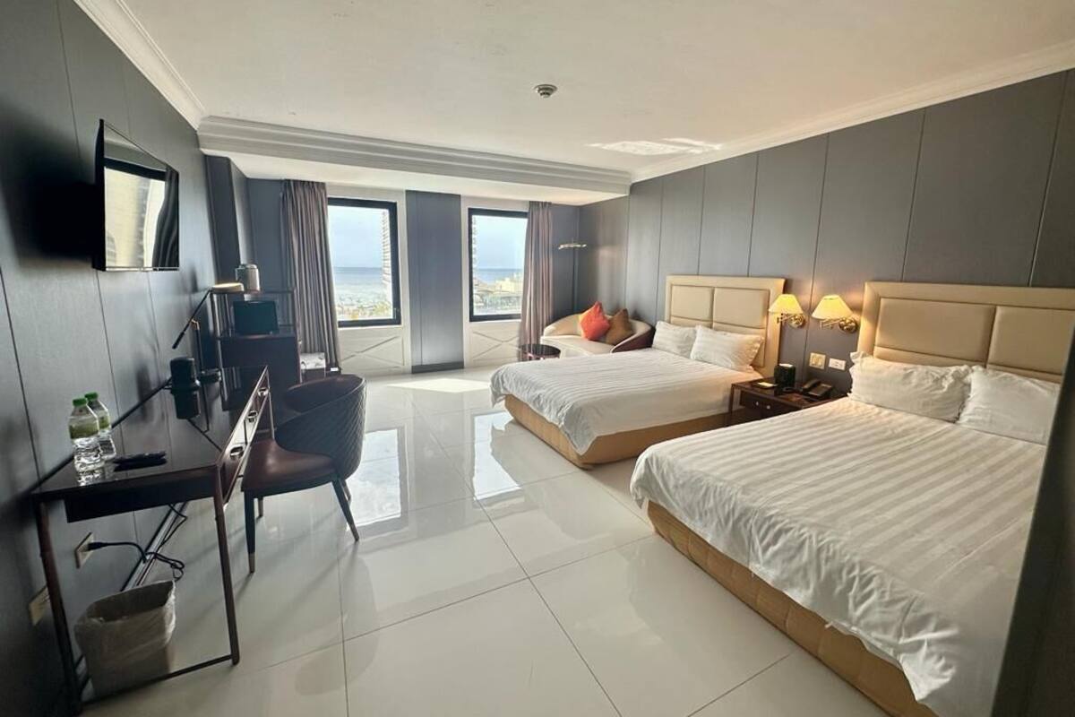 A hotel room with two beds, a desk, a chair, a TV, and a view of the sea.