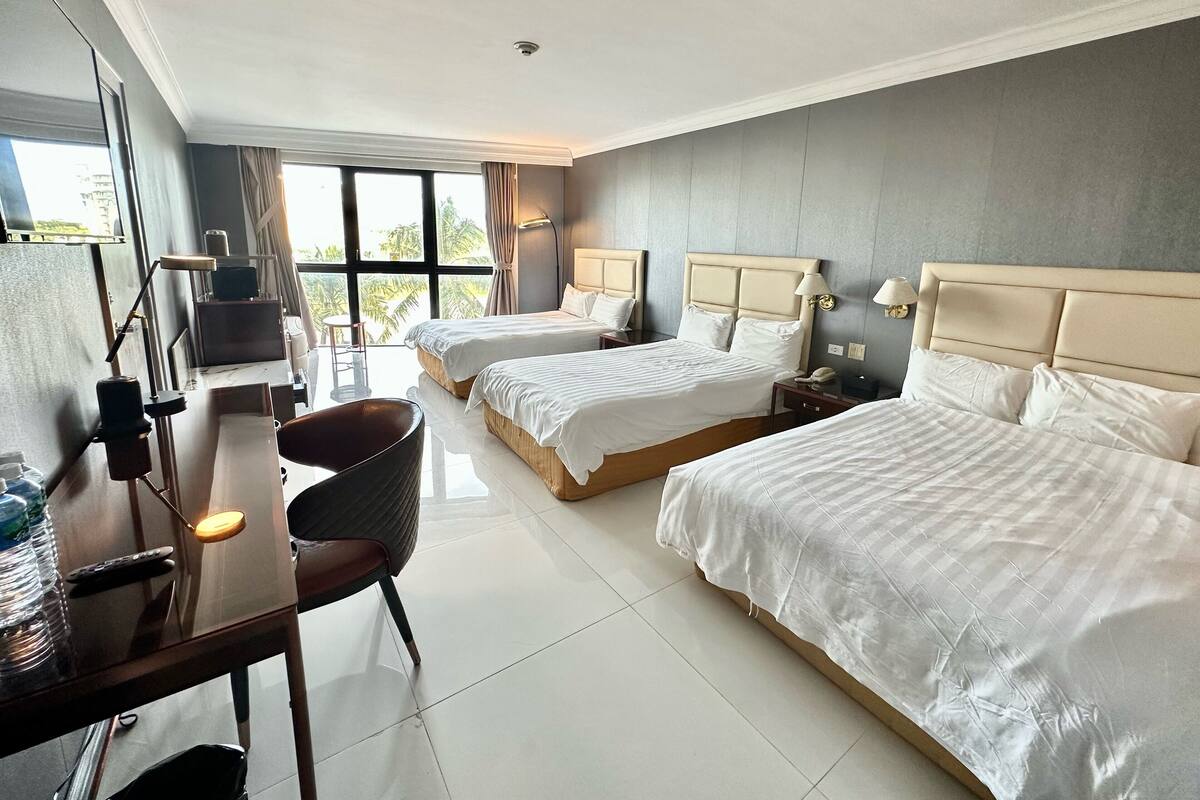 A hotel room with two beds, a desk with a chair, and a large window with a view of palm trees.