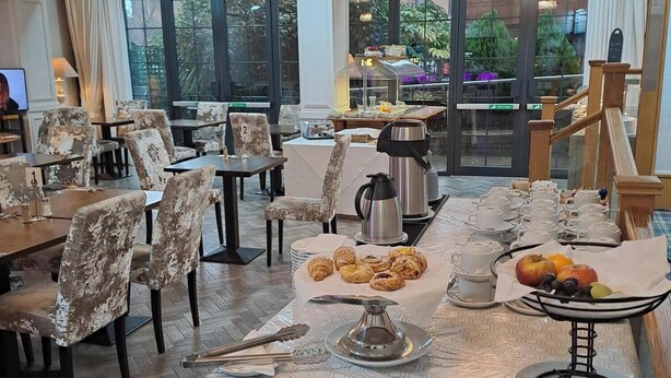 Un espace repas avec des tables préparées pour le petit-déjeuner, comprenant des viennoiseries et des fruits, dans un hôtel moderne doté de grandes fenêtres et d’un lustre.