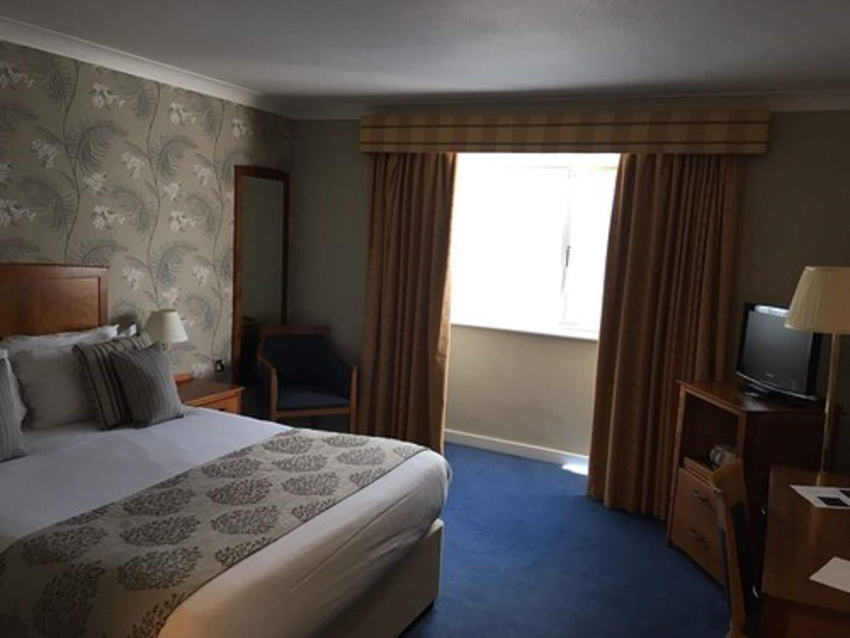 A hotel room with a large bed, a chair, a desk with a television, and a window with curtains.