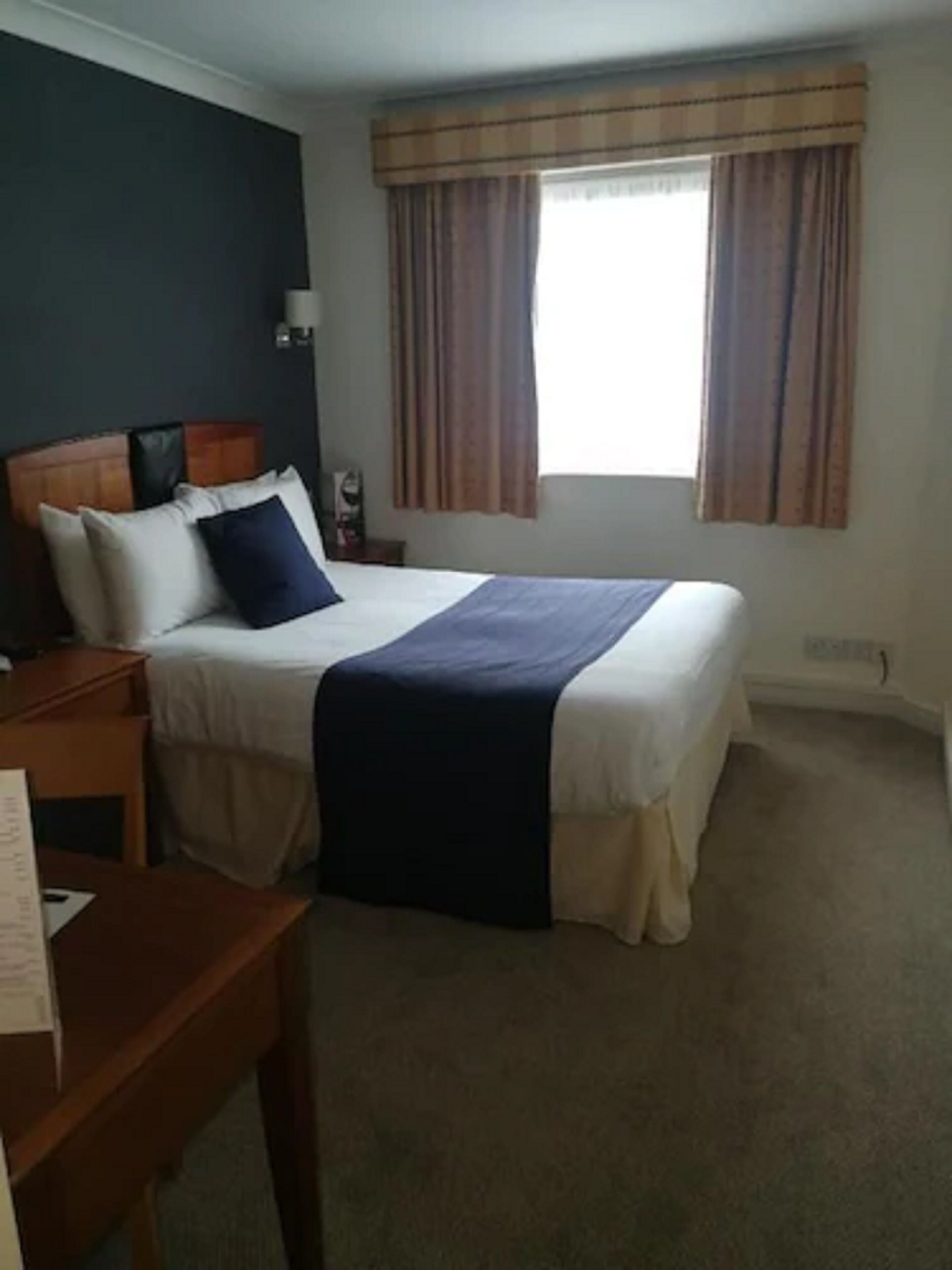 Una habitación de hotel con cama, mesita de noche, escritorio y ventana con cortinas.