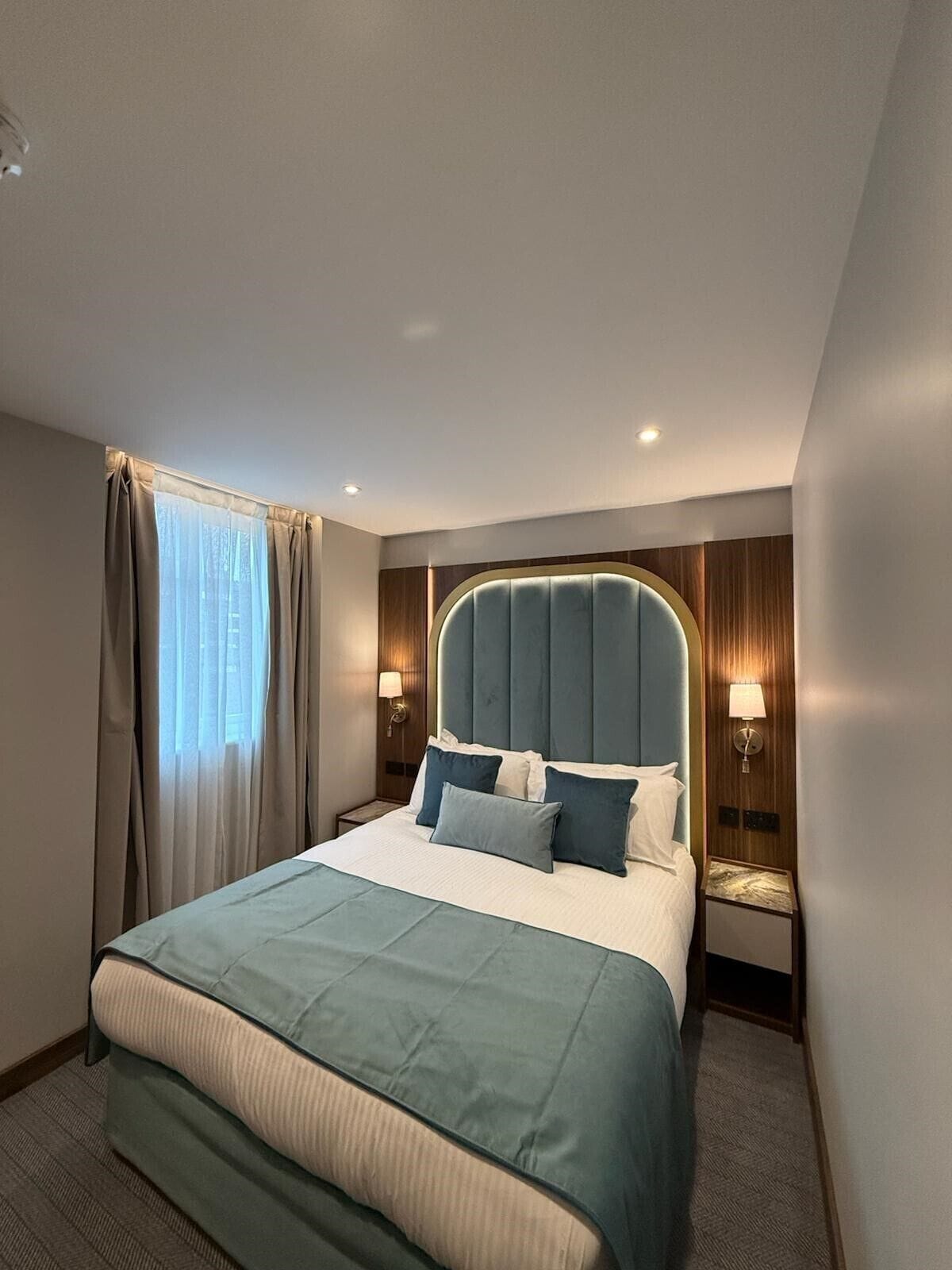 Habitación de hotel con una cama grande, dos mesitas de noche con lámparas, una ventana con cortinas y un cabecero fijado a la pared.