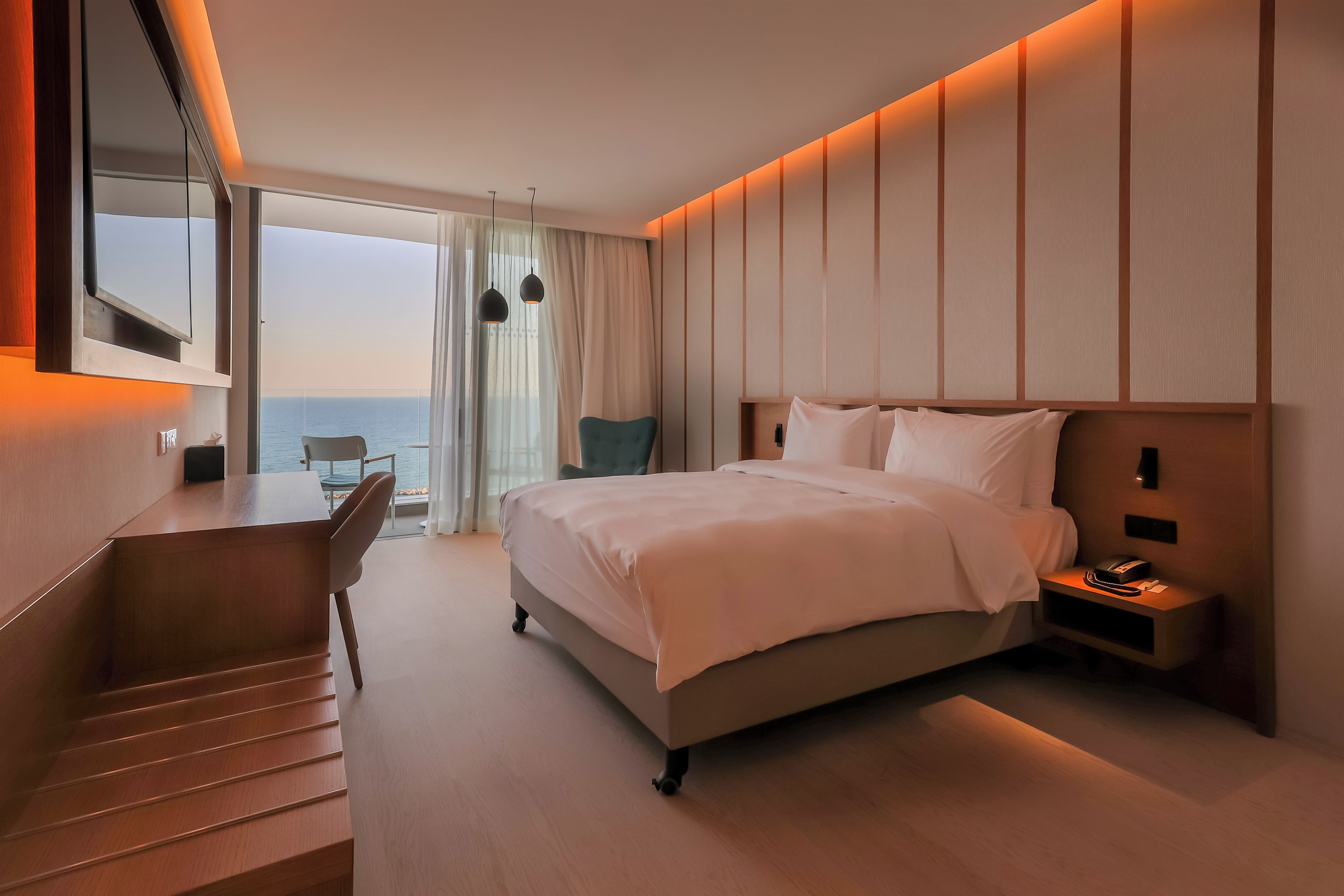 A modern hotel room with a large bed, a desk with a chair, a small table, and a view of the sea through large windows.