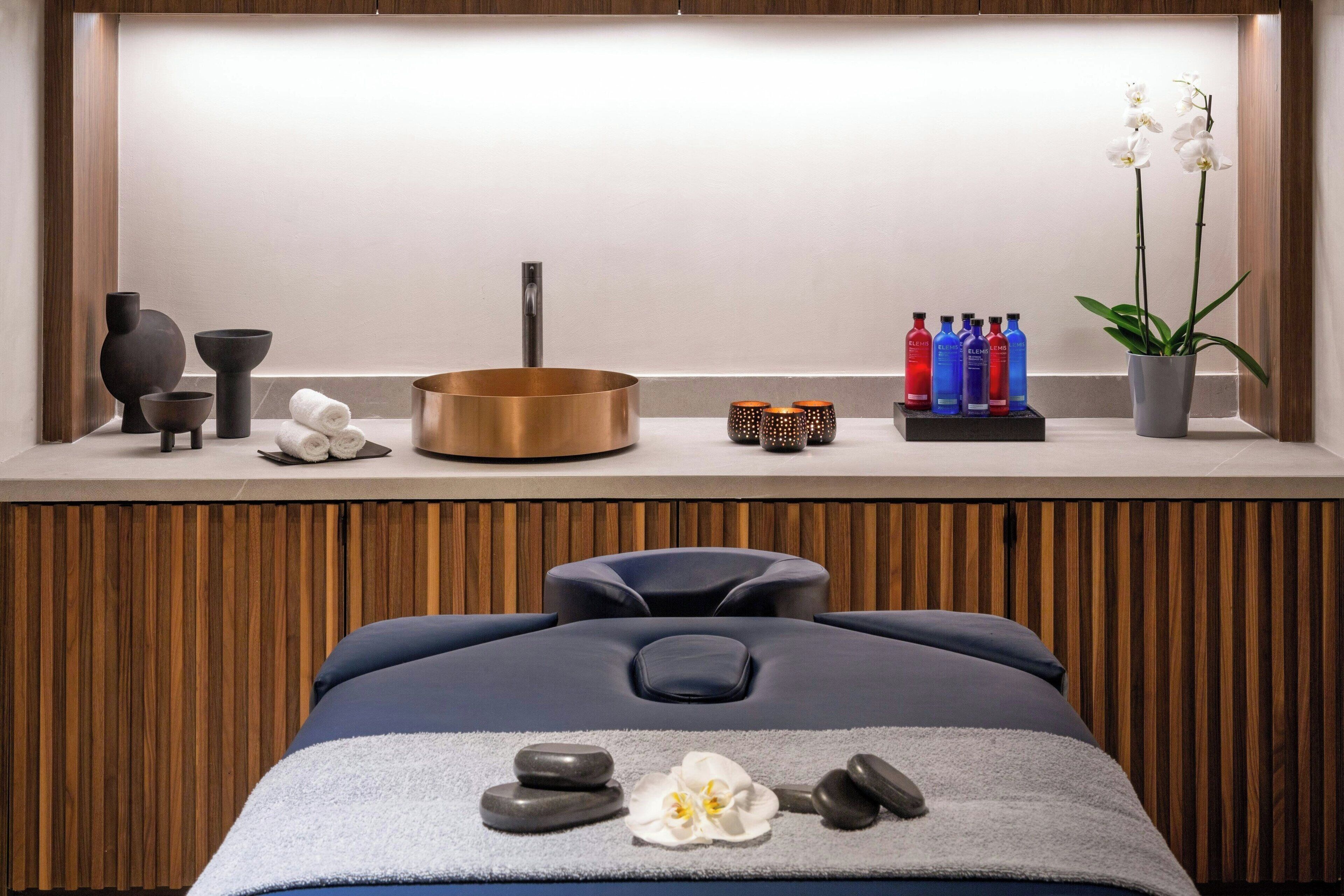 Un moderno spa con una mesa de masaje, un panel de pared de madera, un lavamanos, una orquídea en maceta y botellas de agua.