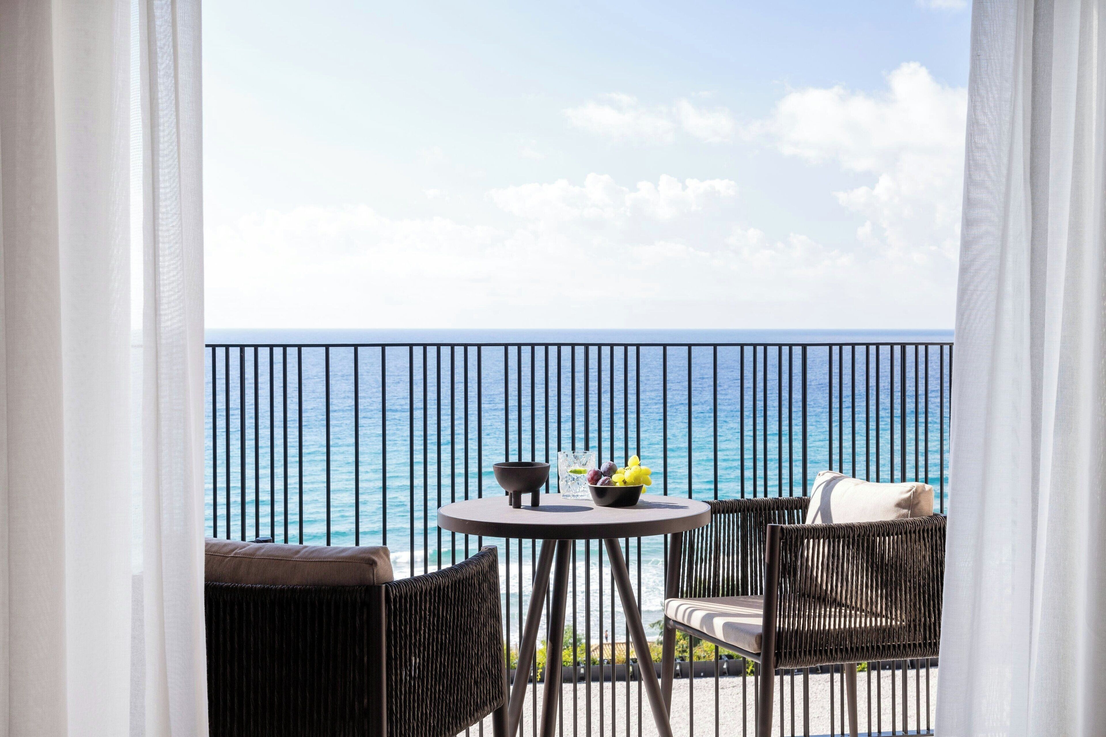 Een balkon met uitzicht op zee, met een ronde tafel, een schaal fruit en een glas water.
