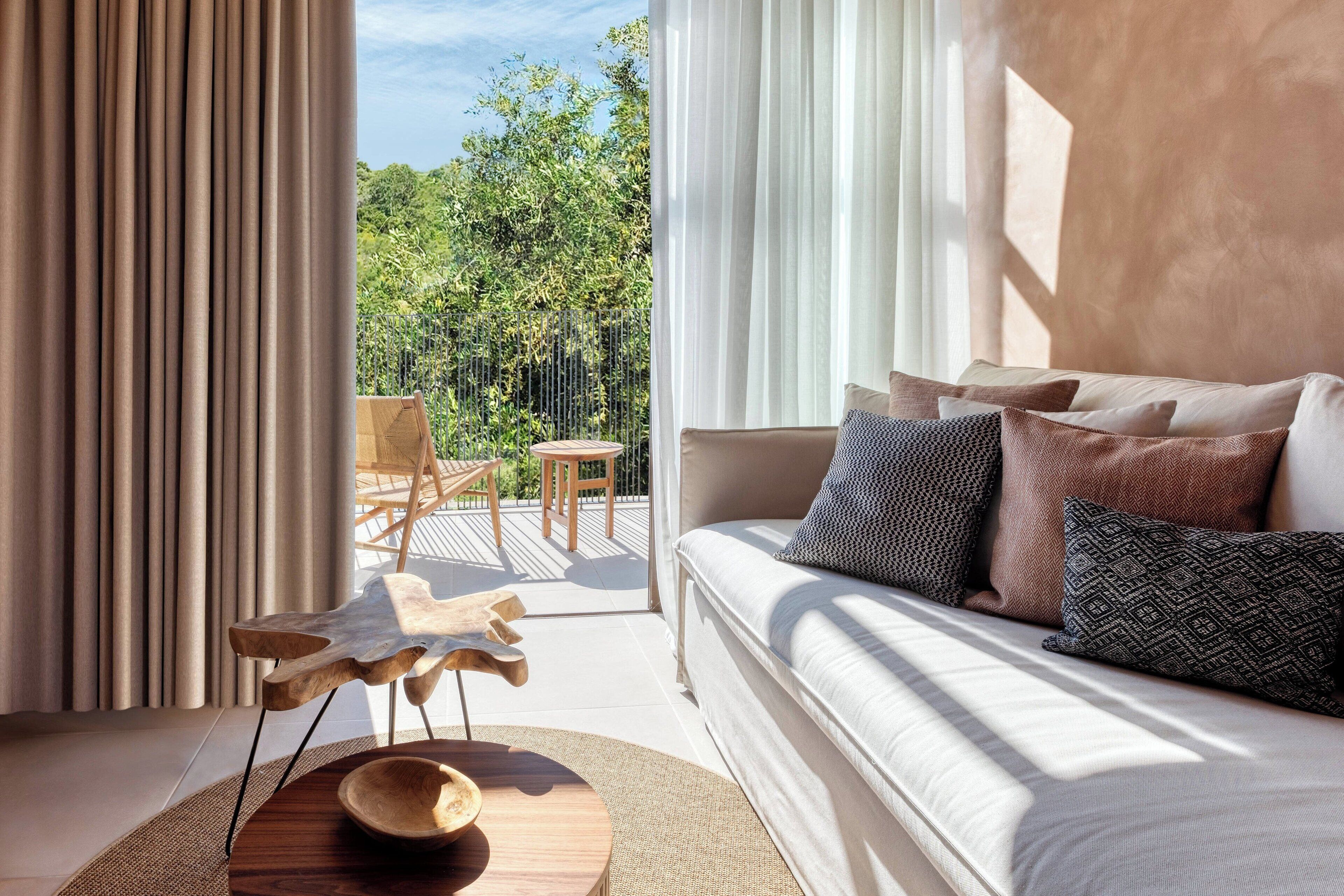 Un salon moderne avec un canapé, des coussins, une petite table en bois et une vue sur une terrasse extérieure et la verdure.