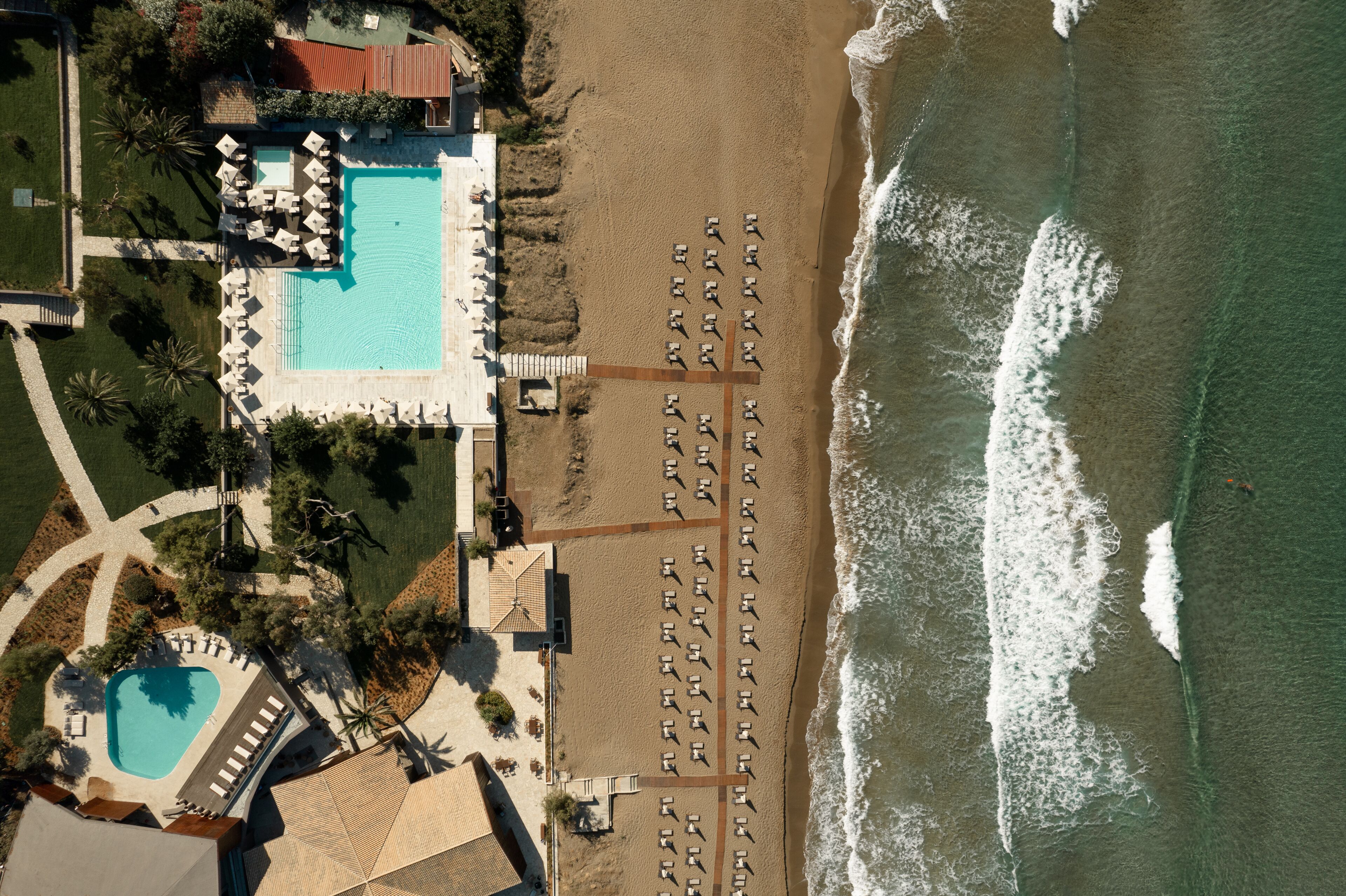 An aerial view of a resort with a swimming pool, beach with sun loungers, and a beachfront view.