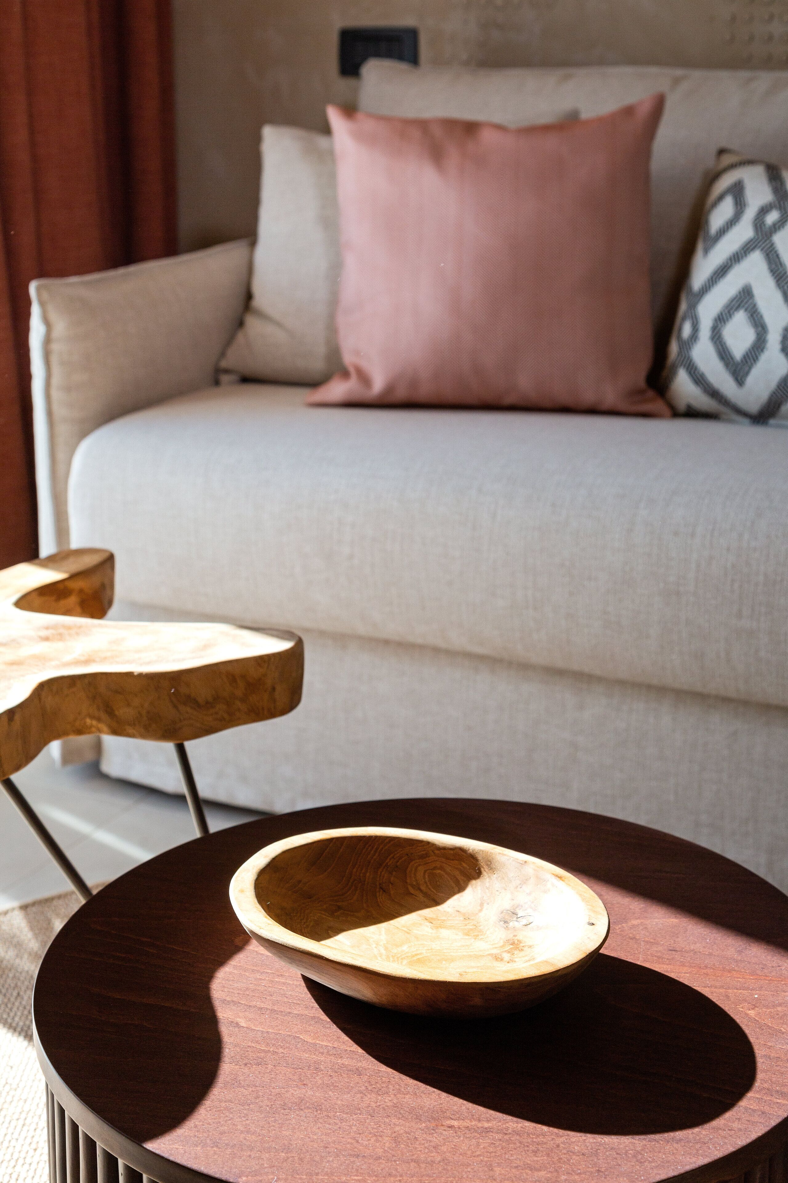 A round wooden table with a bowl on it, a sofa with cushions, and a patterned pillow in a living room.